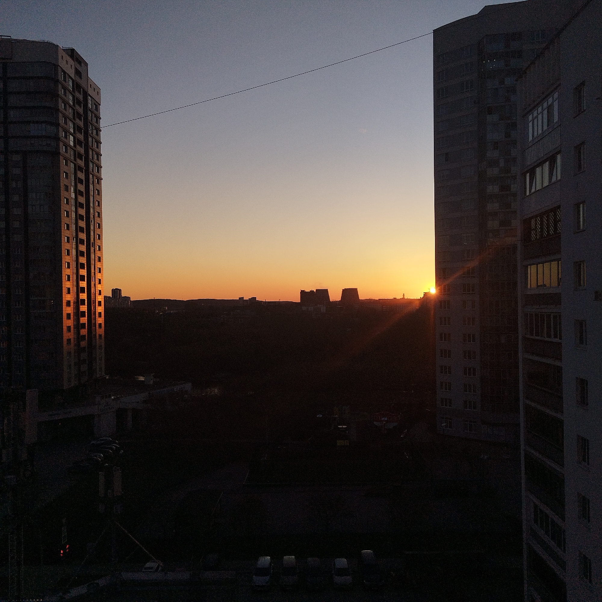 The image captures an urban landscape at either sunrise or sunset, dominated by two tall residential buildings framing a distant horizon. On the left, a multi-story building stands vertically, its windows reflecting the warm, golden light of the low sun. On the right, another large building, appearing slightly wider, partially obscures the bright sun, which casts a strong beam of light diagonally across the scene and illuminates the side of the building with a warm glow.
Between the two buildings, in the middle ground, is a dark, silhouetted landscape of what appears to be trees and smaller structures, stretching towards the horizon where more distant buildings are faintly visible against the bright sky. The sky transitions from a warm, intense orange-yellow near the horizon to a softer, desaturated blue-grey overhead. A thin, dark cable or wire runs horizontally across the upper part of the sky, connecting or passing between the two main buildings.
The foreground and the lower sections of the image are plunged into deep shadow, rendering details almost imperceptible. However, faint outlines of parked cars and possibly a street or courtyard can be discerned in the extreme bottom. The overall impression is one of tranquility and the stark beauty of natural light interacting with man-made structures in an urban setting. No discernible text is visible in the image.