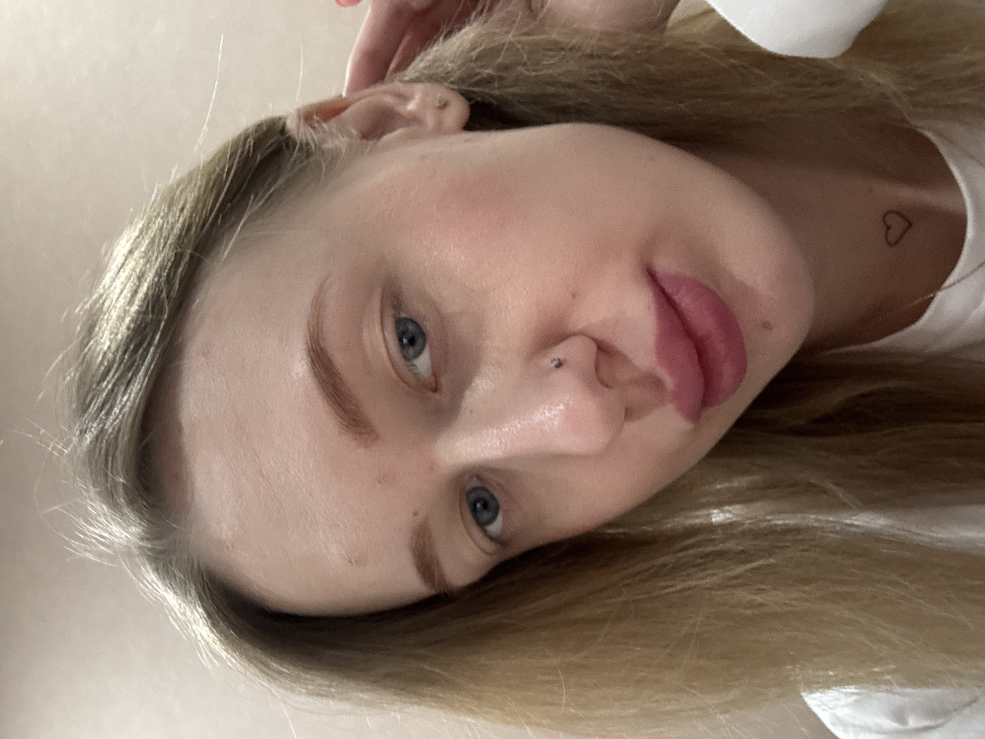 A close-up, angled headshot of a fair-skinned woman with light blonde hair and blue eyes. She has a nose piercing and a small heart tattoo on her collarbone. The background is a plain, light-colored wall. The location is not determinable from the image.