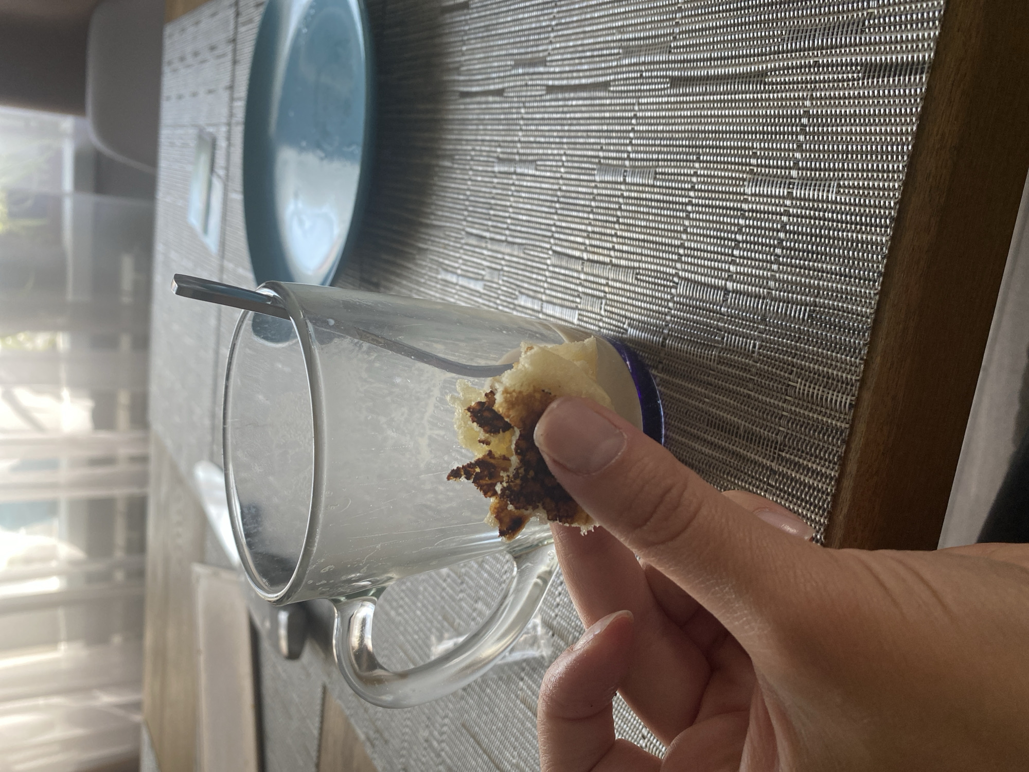 A person's hand holds a piece of a sweet pastry, appearing to be cake or muffin, towards a clear glass mug on a placemat. The mug contains a small amount of liquid and a spoon. In the background, a blue plate sits on the placemat, and a window with sheer curtains is visible, suggesting an indoor setting, possibly a dining area. The lighting appears natural and diffused.