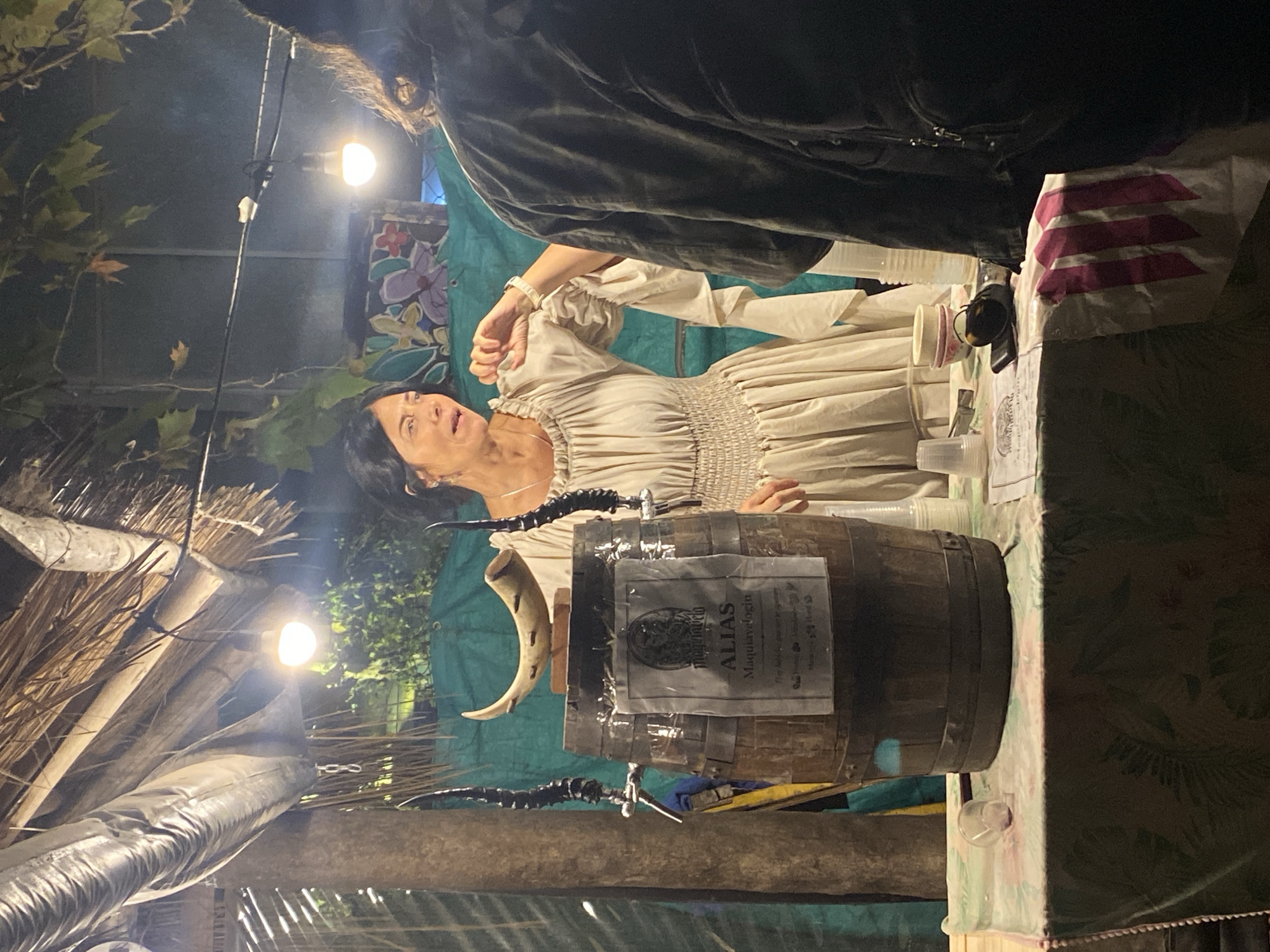 The image captures a scene at what appears to be an outdoor market or festival, possibly at night, as evidenced by the artificial lighting. A woman wearing a period-style dress with ruffled sleeves stands behind a table or counter. On the counter is a small barrel labeled "ALIAS Maquiavelogia" with a spigot and a horn-shaped object attached. The setting might be the city of Buenos Aires, Argentina, given the reference to "Maquiavelogia," which sounds like a Spanish/Italian concept. An individual is only partially visible from the back on the right side. The area around the vendor is adorned with straw-like roofing, suggesting an outdoor event. Overall, the scene suggests a historical or cultural event.