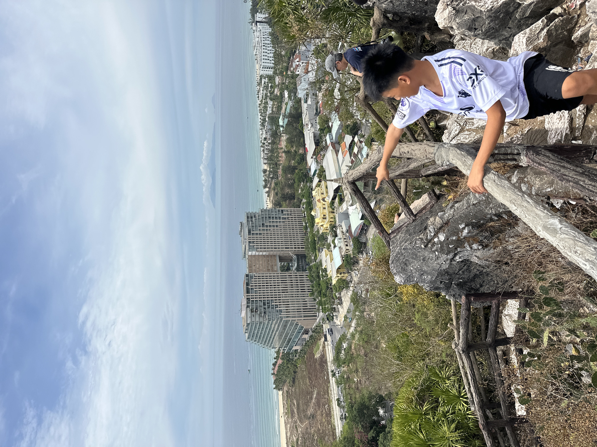 A young boy in a white t-shirt and black shorts is standing on a rocky outcrop, pointing towards the sea. He is on the right side of the frame, his back partially to the viewer. Behind him, a man wearing a cap and dark shirt stands with a green camera bag slung over his shoulder. They are positioned on a wooden railing overlooking a city with a beach and ocean in the distance.
The city in the background features several modern buildings, including a distinctive, curved hotel with a reflective facade, and various other low-rise structures. The beach is visible with the turquoise water of the sea meeting the sandy shore. The sky is partly cloudy, with a hazy horizon.
The foreground is dominated by rocks and rugged terrain, with a rustic wooden fence in place, suggesting a viewpoint or park area. The vegetation includes some palm-like plants and dry grasses. The scene appears to be a scenic overlook in Đà Nẵng, Vietnam, likely offering a panoramic view of the coastline and urban development.