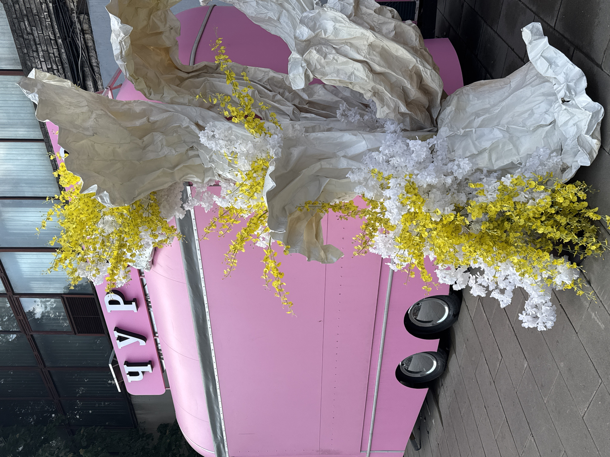 A vibrant pink food truck, possibly branded with Cyrillic lettering, is parked on a stone pavement in front of a building with large industrial windows. The truck is adorned with a dramatic floral and paper art installation. Clusters of delicate yellow and white artificial flowers cascade down, intertwined with large, crumpled white paper structures resembling abstract sculptures. The scene appears to be outdoors, with a hint of urban decay in the background building. The overall impression is one of playful, artistic decoration contrasting with a utilitarian urban setting, potentially in the city of Saint Petersburg, Russia, given the Cyrillic lettering.