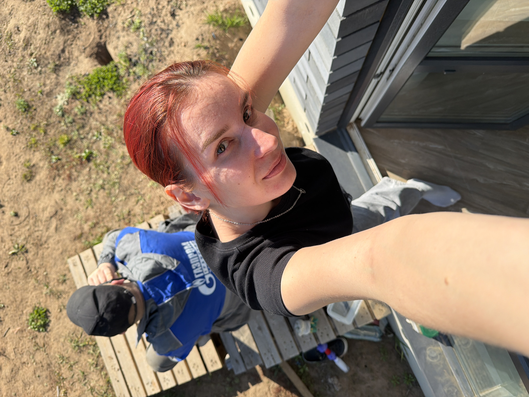 A high-angle photograph taken in Chaikovsky, Russia shows a person with short red hair taking a selfie. The subject is wearing a black top and a silver necklace. Behind them and slightly to the left, a man in a blue and gray work uniform and a black cap is lying on a wooden pallet. The word "GAZPROM" is partially visible on the uniform. The background consists of a building exterior with gray siding and a window, along with a patch of bare earth with some sparse vegetation. The photo is taken in daylight with strong shadows.