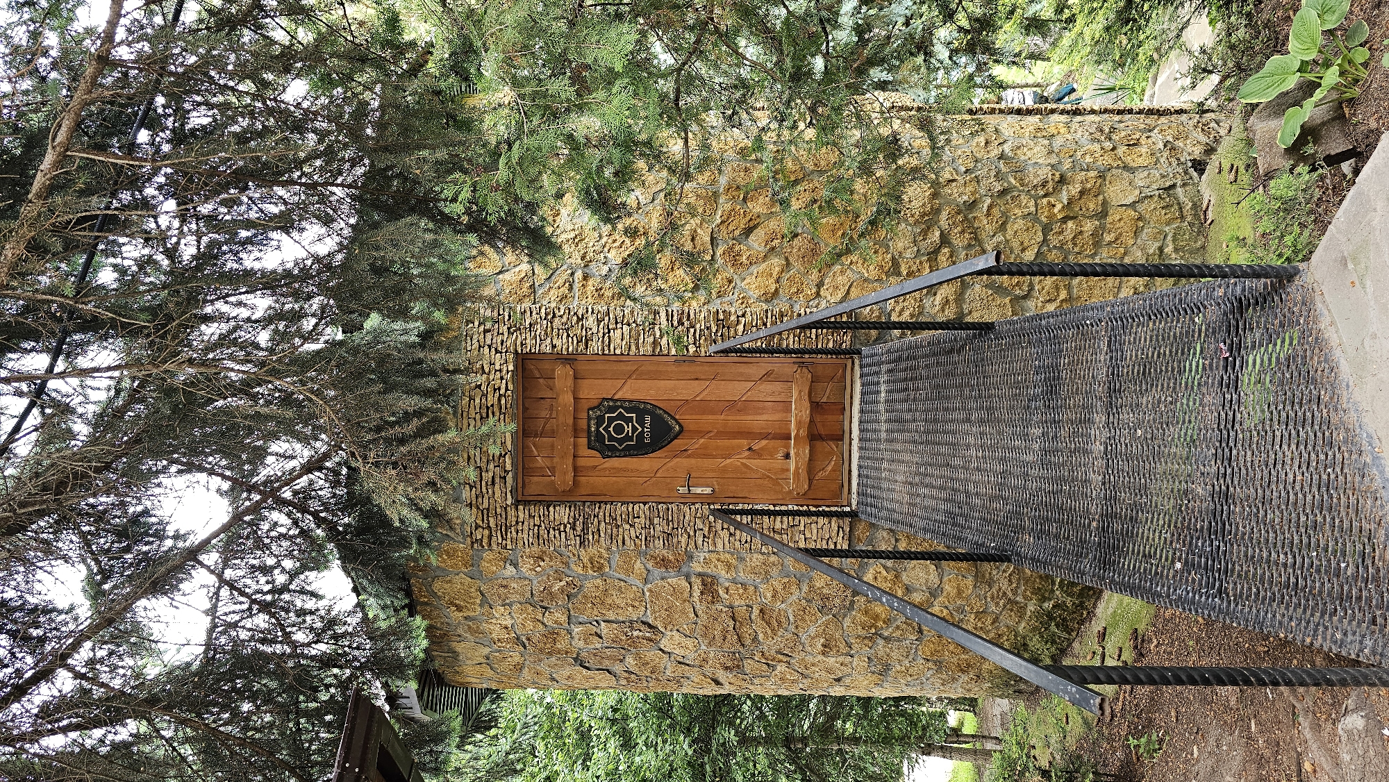 A weathered wooden door is set into a round tower constructed of rough-hewn, honey-colored stones. The door is adorned with a black, shield-shaped emblem featuring a circular design and the Cyrillic word "БОТАШ" beneath it. A metal ramp with black railings leads up to the entrance. The structure is surrounded by lush green trees and foliage, with a dense canopy of pine branches overhead partially obscuring the sky. The scene suggests a rustic, possibly historical or themed, building within a natural setting, likely in the city of Krasnyy Kurgan, Russia.