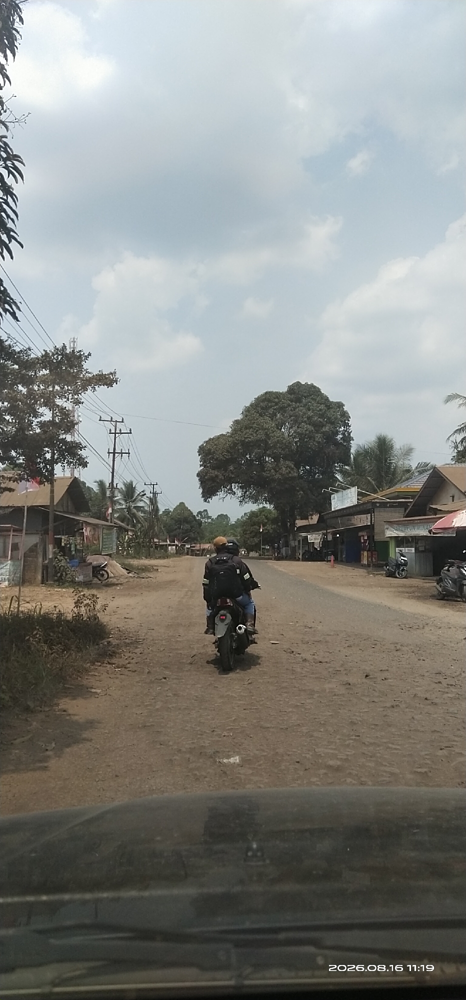 This is a slightly low-angle, full-shot, vertical image taken from the perspective of a vehicle, looking down a dirt road in Jaweten, Indonesia. Two individuals, a driver and a passenger, are riding a motorcycle away from the camera. The driver is wearing a light brown helmet and a black jacket with reflective stripes. The passenger is wearing a black backpack. The motorcycle is gray and silver. The road is unpaved and appears dusty with some debris scattered on it. On either side of the road are a few low-lying buildings, some with simple wooden structures and corrugated metal roofs. There are trees and utility poles lining the road. In the distance, the road curves to the right and appears to lead into a more densely vegetated area. The sky is partly cloudy with a light blue hue. The timestamp "2026.08.16 11:19" is visible in the lower right corner of the image, possibly indicating the date and time the photograph was taken.