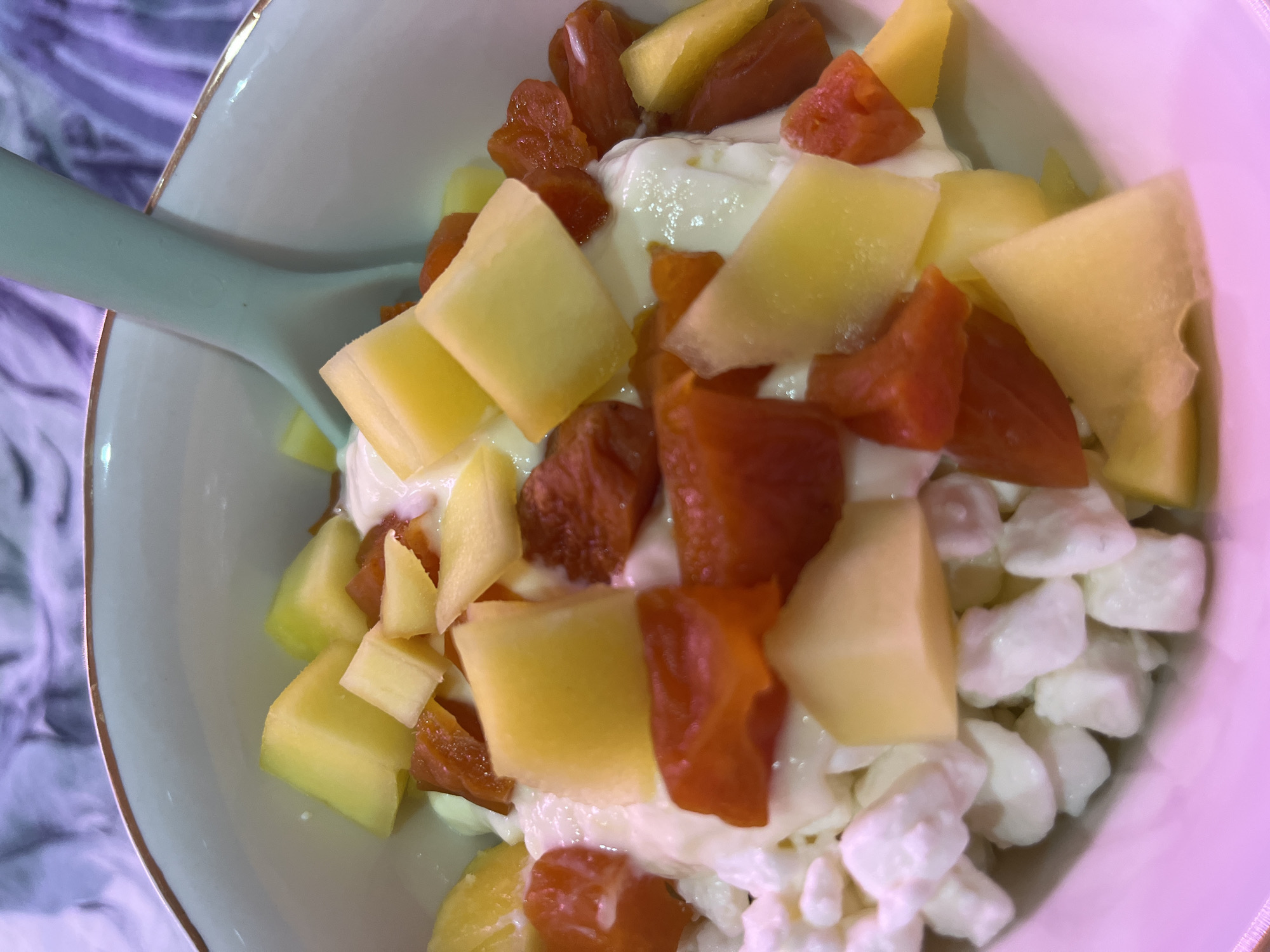 A close-up, high-angle shot captures a bowl filled with a meal or snack, primarily consisting of fruit and a creamy base. The dish features generous portions of diced light yellow fruit, likely melon such as cantaloupe or honeydew, mixed with smaller, darker orange-red pieces of fruit, possibly dried apricots or papaya. These fruits are combined with a white, lumpy, creamy substance, resembling cottage cheese or yogurt, which fills the bottom of the bowl.
A light blue-green spoon rests within the bowl, partially submerged in the food, indicating that the meal is prepared and ready for consumption. The bowl itself is light-colored, possibly off-white or pale blue, and features a subtle gold rim. The background is softly blurred, revealing a patterned fabric in shades of purple, blue, and white, likely a tablecloth or blanket. No people are visible, and there is no discernible text in the image. The setting appears to be indoors, in a general home environment.