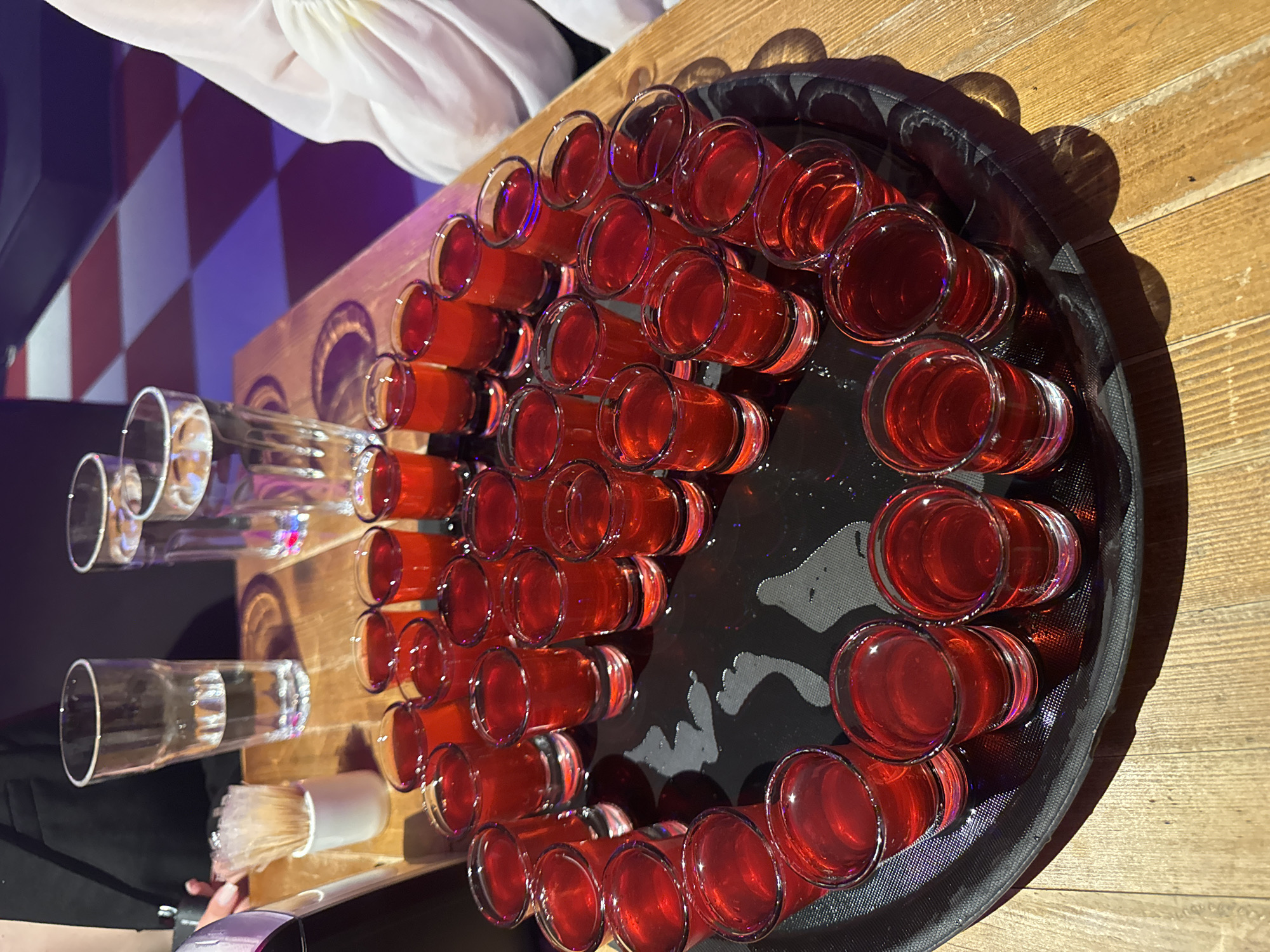 The image shows a close-up, high-angle view of a black tray filled with numerous small glasses of red liquid, likely shots of alcohol. The tray is placed on a wooden surface. In the background, there are taller glasses of clear liquid and what appears to be a patterned floor with red and blue squares, suggesting a bar or club setting. A person's arm, wearing a black jacket, is visible on the left side of the image, with a hand reaching towards some toothpicks in a small white holder. Another person, partially visible on the right, is wearing a white puffy garment. The lighting is dim with some colorful reflections on the glasses, indicating artificial lighting. There is no discernible information in the image to suggest a specific location in the city of Perm, Russia.