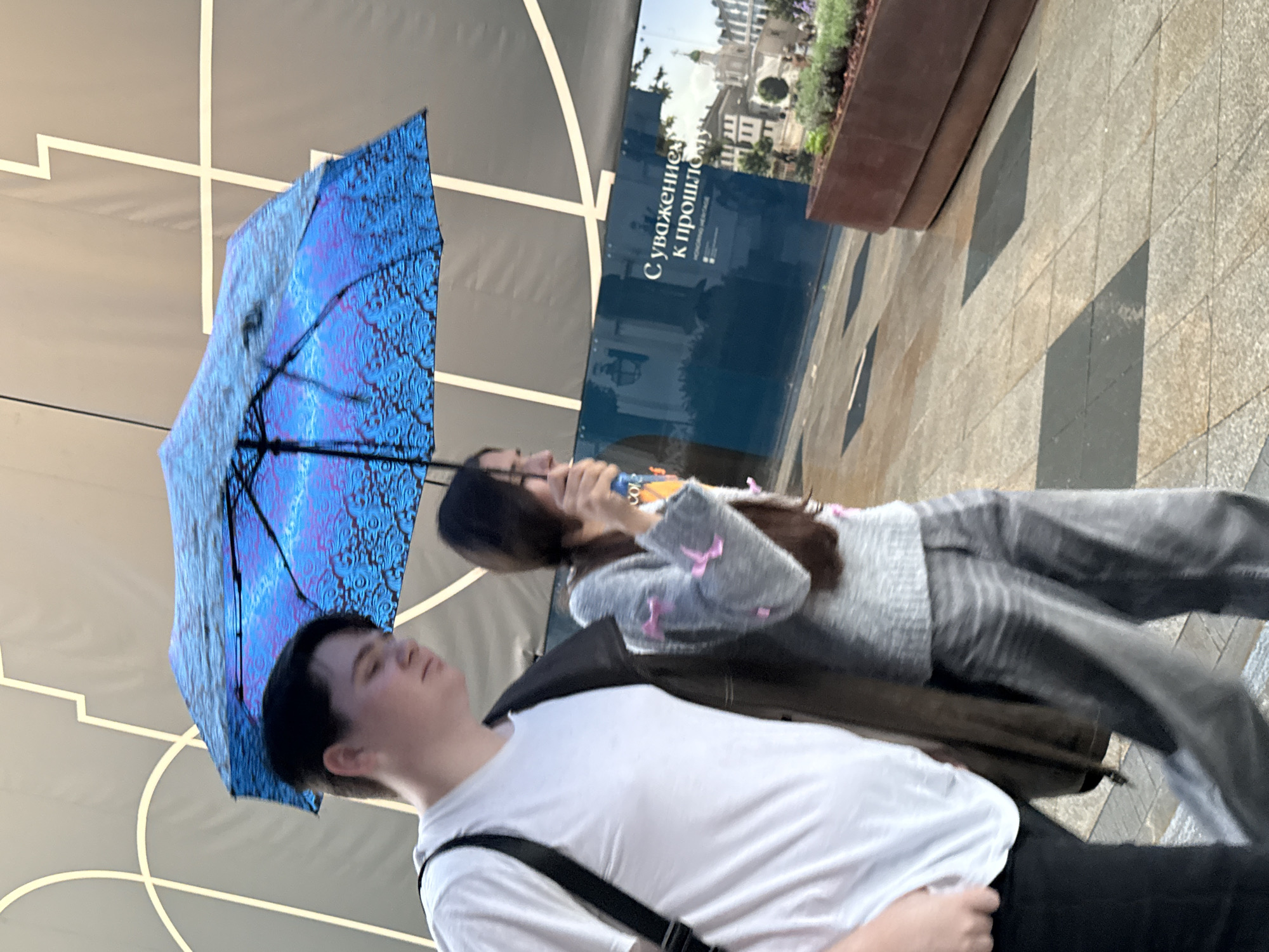 Two individuals are walking under a blue umbrella with a patterned design, suggesting it might be raining or to provide shade from the sun. The person on the left is wearing a white t-shirt and black pants, with a black strap across their chest. They have light-colored skin and dark hair. The person on the right appears to be wearing a grey sweater with light pink embellishments and grey pants. They have long dark hair and are holding the umbrella. Behind them, there is a large blue sign with white text that appears to be in Russian. Part of the text reads "С уважением к прошлому" (With respect to the past). The background also shows a building or architectural element with white lines on a grey surface. In the foreground, there is a paved walkway with square stone tiles, and a raised planter box with greenery. The lighting suggests it is daytime. This scene is likely taking place outdoors in an urban environment.
The location in the city of Moscow, Russia is the Mosgortans Exhibition Center on the bank of the Moskva River, in front of the building of the State Duma.