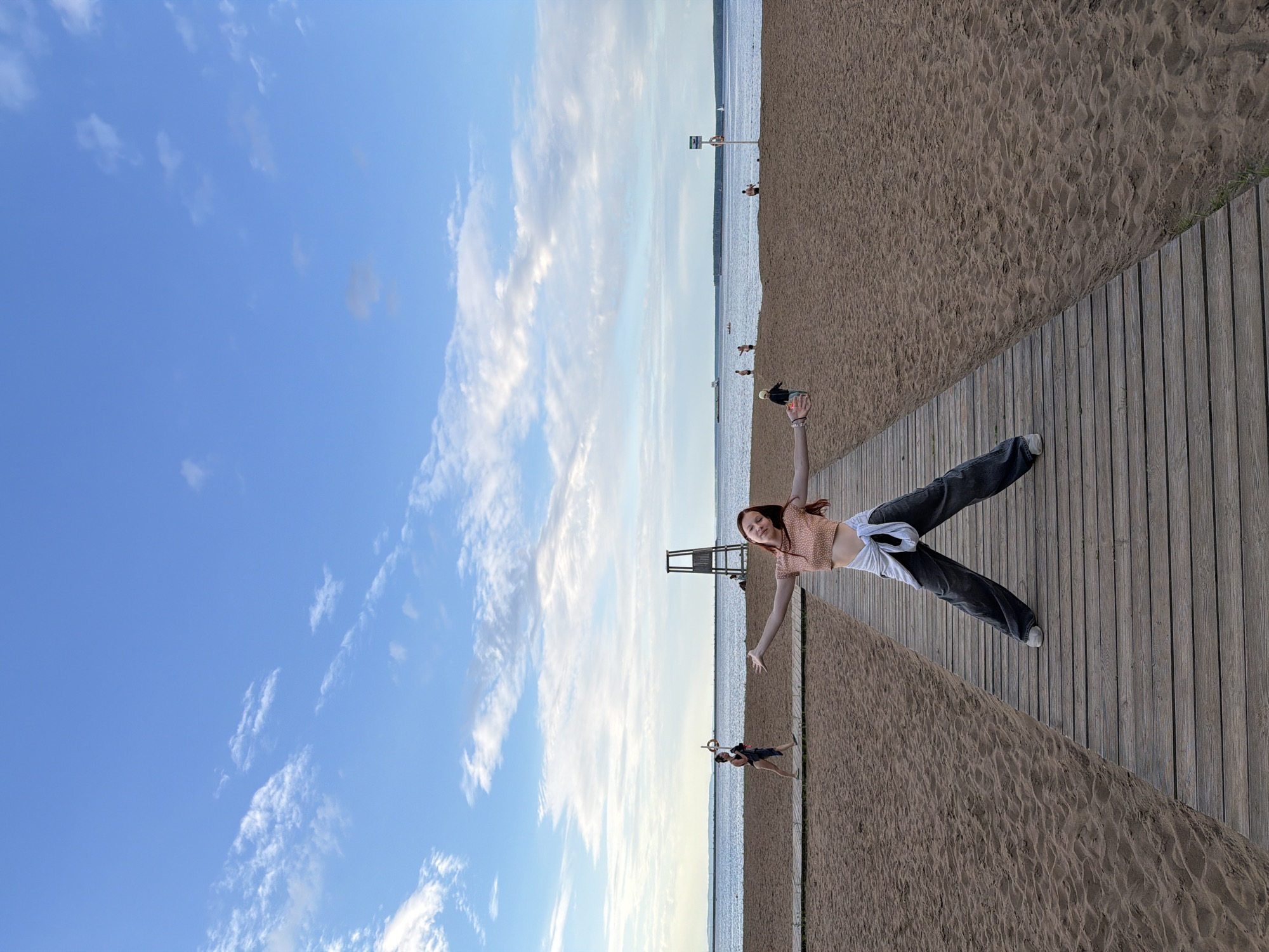 The image depicts a young woman with red hair standing on a wooden boardwalk on a beach. She is wearing a peach-colored crop top and dark, wide-leg jeans, with a white shirt tied around her waist. Her arms are outstretched to the sides, and she is looking at the camera with a smile. The boardwalk extends into the distance, with sand on either side. In the background, a body of water is visible, with a few people and a lifeguard tower on the beach. The sky is blue with scattered clouds. The location appears to be a recreational beach area.