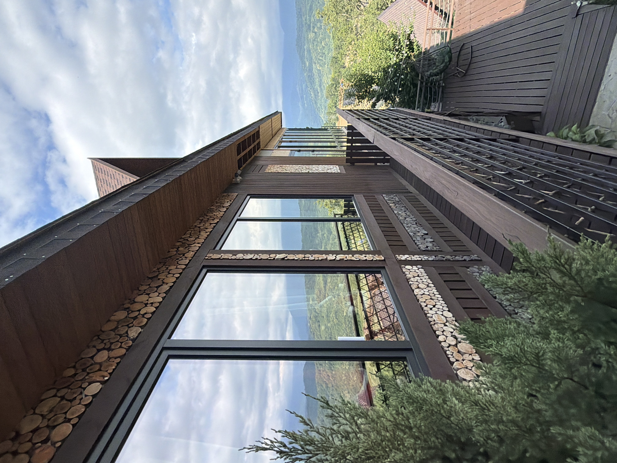 This is a slightly high-angle, eye-level outdoor shot of a modern wooden building with large windows, situated on a hillside with a view of a valley and distant mountains. The building's exterior is decorated with rows of circular wood slices. A dark wooden balcony with a metal railing runs along the side of the building, overlooking a wooden deck. In the foreground, a portion of a green evergreen tree is visible. The sky is partly cloudy. The scene appears to be located in Nizhnyaya Shilovka, Russia, given the architectural style and natural landscape. There are no people or animals visible in the image.