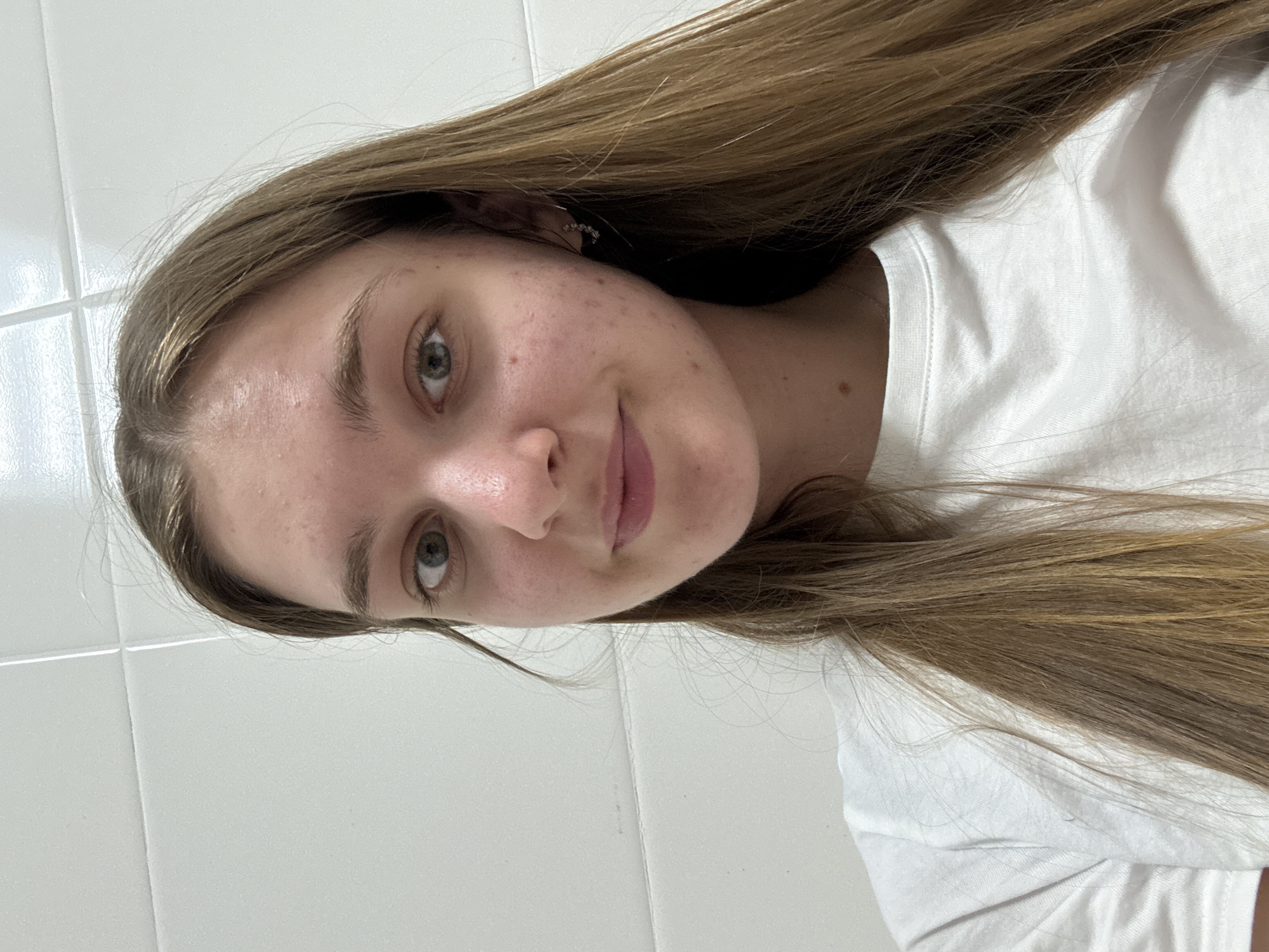 A young woman with light brown hair and fair skin is posing for a selfie. She is wearing a white T-shirt. The background appears to be a white tiled wall. There is no indication of this image being taken in Perm, Russia.