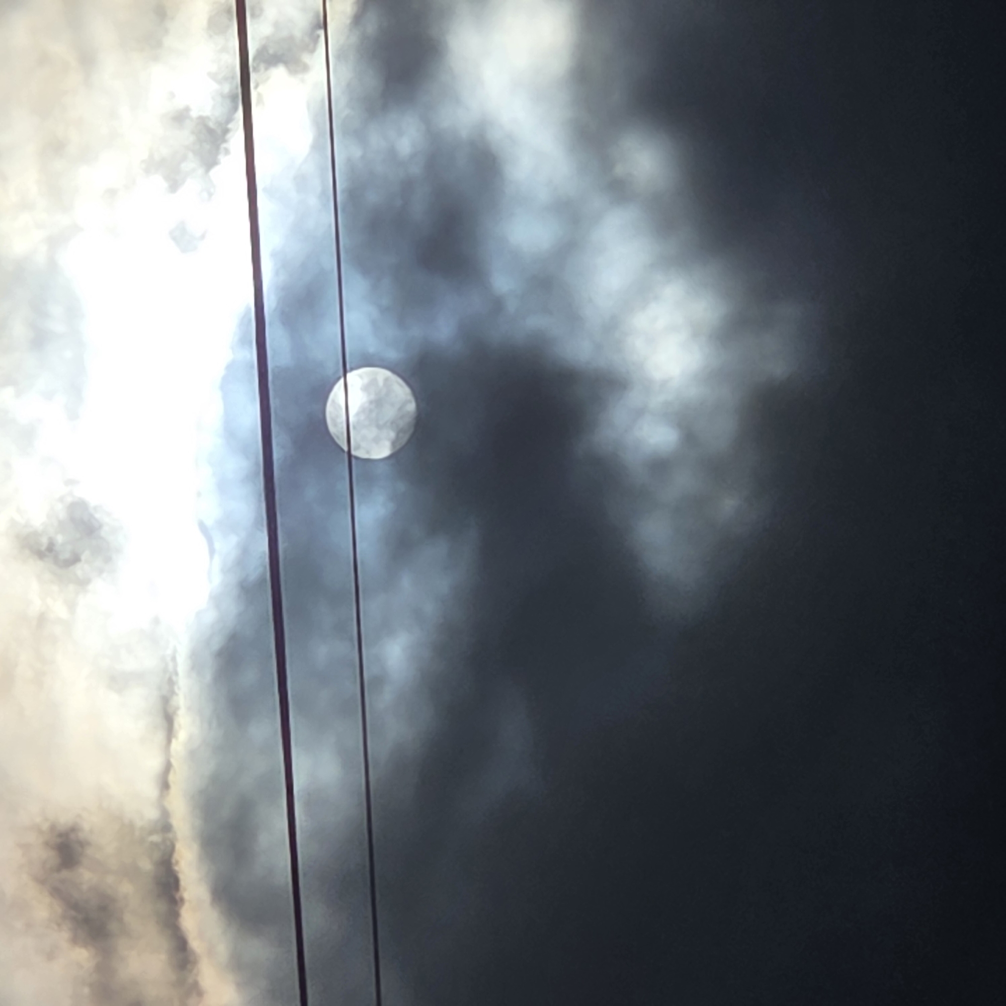 The image shows a cloudy sky with the sun partially obscured by dark clouds. A round, bright object, likely the sun or moon, is visible through a gap in the clouds. Two horizontal dark lines, which appear to be power lines, cut across the frame. The foreground is dominated by dark, ominous clouds, while the background shows lighter, more diffuse clouds and the bright celestial body. The scene is foreboding and dramatic, with the light struggling to break through the heavy cloud cover. The location is identified as Dunnae-myeon, South Korea. There are no people or specific structures visible in the image, only the atmospheric elements and the power lines.