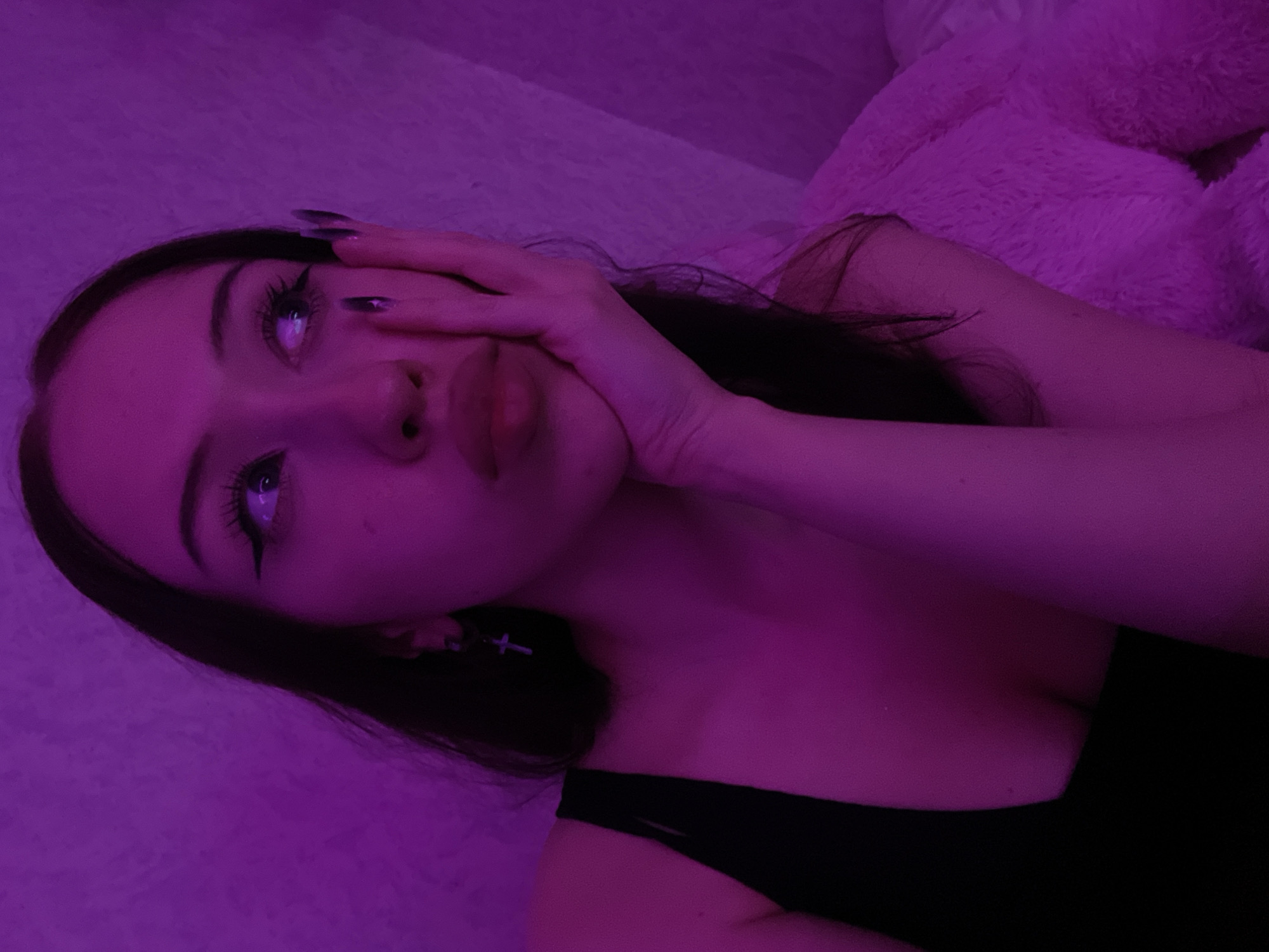 A young woman with long dark hair and winged eyeliner looks to her right with a pout. Her left hand is propped on her cheek, with long dark fingernails. She wears a black sleeveless top and a silver cross earring. The scene is illuminated by purple lighting, creating a moody atmosphere. The background appears to be a textured wall and a fuzzy pink blanket. There is no information in the image that places it in Votkinsk, Russia.