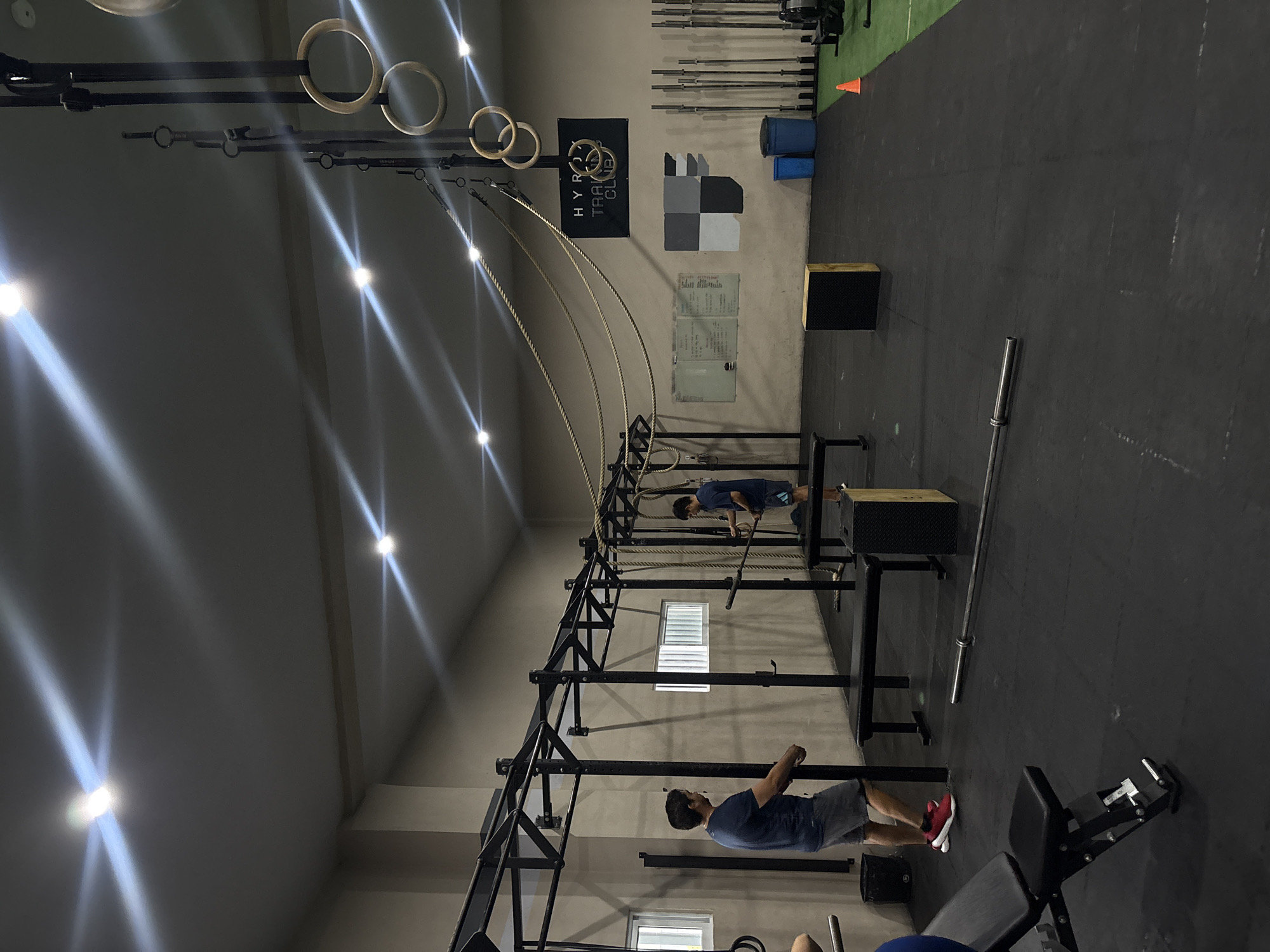 The image depicts the interior of a gym, possibly "HYROX Training Club," as indicated by a banner on the wall. Two men are present. One man, wearing a blue t-shirt and grey shorts, stands facing a squat rack and appears to be preparing for an exercise. The second man, also in a blue t-shirt and grey shorts, is to the right, holding a barbell and engaging in activity near a plyometric box. The gym environment features specialized workout equipment including a squat rack, barbells, gymnastic rings suspended from the ceiling, a plyometric box, weight plates, and weight machines. The floor is covered in black rubber matting. Natural light enters through a window on the left wall, and artificial lighting is provided by overhead fixtures that create prominent light flares. The overall scene suggests a functional training or CrossFit-style gym. The image was taken in San Bernardino Tlaxcalancingo, Mexico.