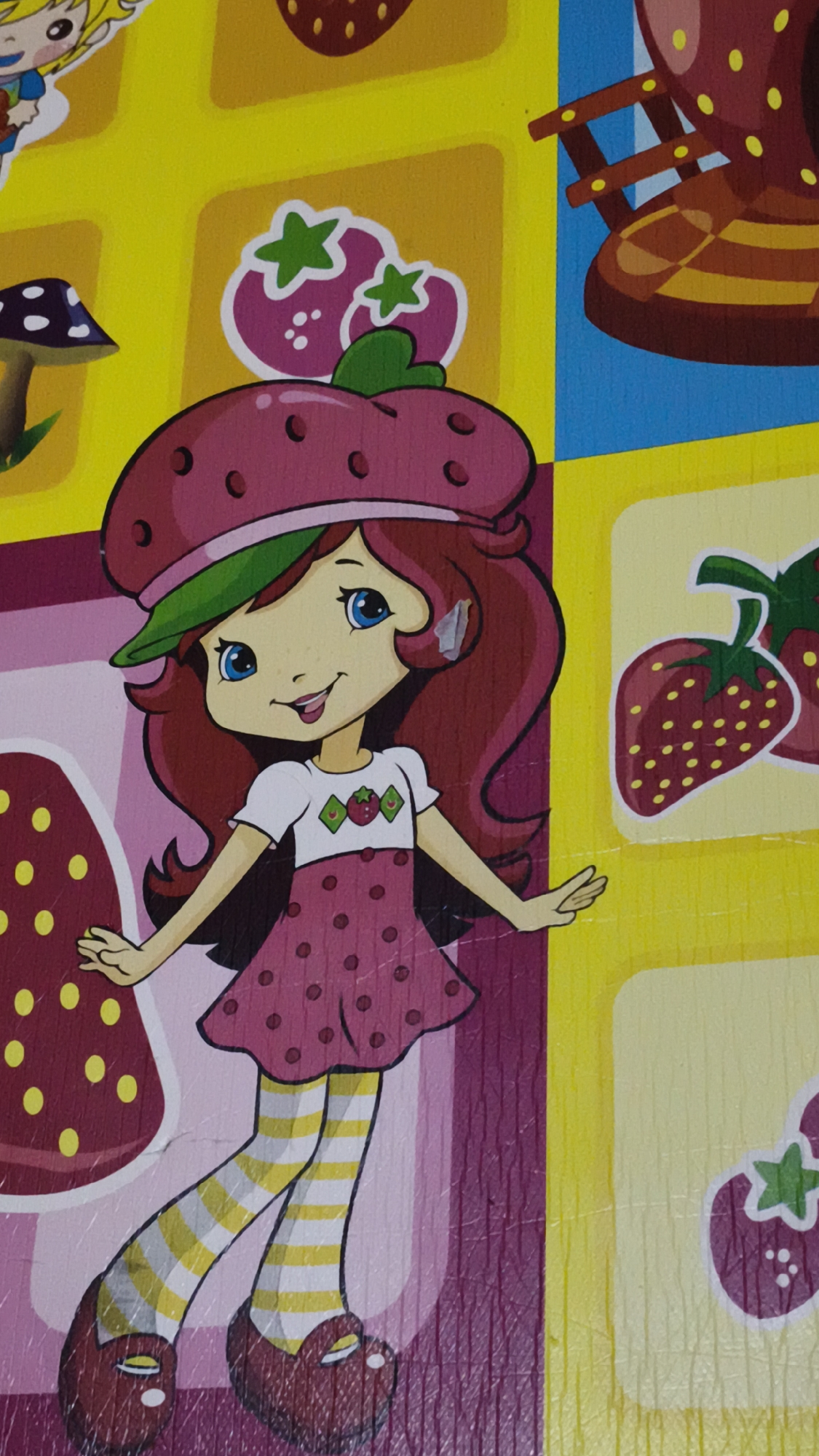 The image is a close-up, vertical, eye-level shot of a cartoon illustration, likely from a children's book or toy. The central subject is a young girl with red hair, large blue eyes, and a wide smile, dressed in a pink polka-dot dress and striped stockings. She is wearing a strawberry-shaped hat with a green brim. To the left of the girl is a mushroom with a purple spotted cap and a brown stem. To the right of the girl are illustrations of strawberries. The background is a grid of brightly colored squares, primarily yellow and blue, with pink accents. The overall style is whimsical and playful. The image is a photograph of a surface, possibly wood, with visible grain and some scuff marks, suggesting it is a manufactured item. There is no indication of Kerep Kidul, Indonesia, or any specific urban environment.