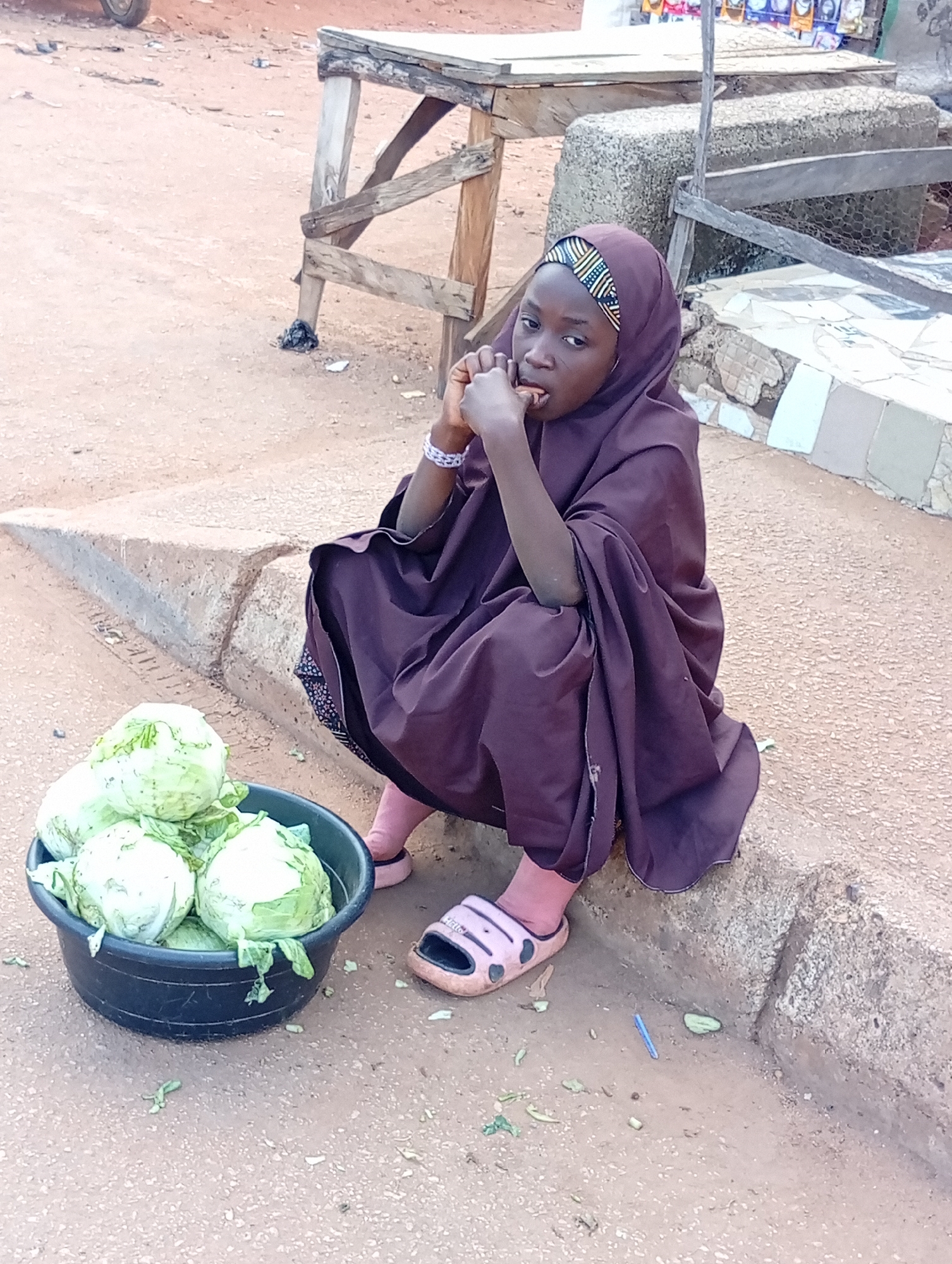In Bukuru, Nigeria, a young girl sits on a concrete curb, her right hand near her mouth and her left arm bent at the elbow. She is dressed in a dark purple hijab and matching garment. A black plastic bowl filled with several heads of lettuce is placed to her left. Pink sandals are on her feet. The ground is dusty and appears to be a dirt road. In the background, a wooden market stall with rough-hewn planks and supports is visible. Further in the background, there are some items for sale on shelves. The overall scene suggests a market or roadside vendor environment.