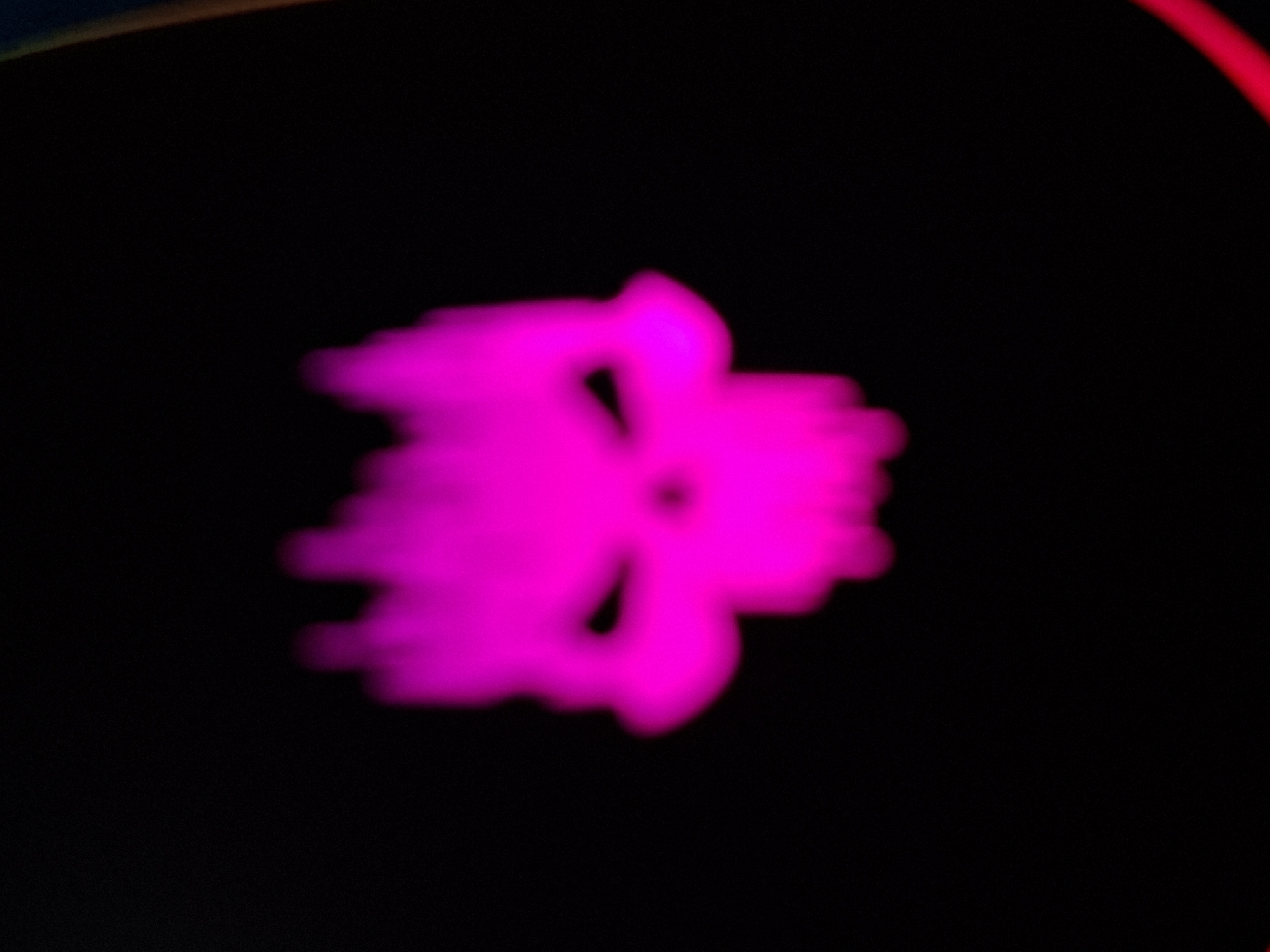 This is a close-up, out-of-focus shot of a bright pink, abstract shape against a black background. The shape appears to have two distinct lobes or sections that are connected in the center, with some wispy, trailing elements. The overall impression is of something organic or fluid, perhaps like a stylized flower, a ghostly figure, or a light effect. There are also hints of red and yellow light in the upper left corner of the image, suggesting the presence of other light sources or reflections. The blurriness of the image makes it difficult to discern precise details or to identify specific objects, but the dominant feature is the vibrant pink color and the energetic, somewhat amorphous form. The darkness of the background intensifies the glow of the pink shape, creating a dramatic and mysterious effect. No people, text, or discernible setting are visible.