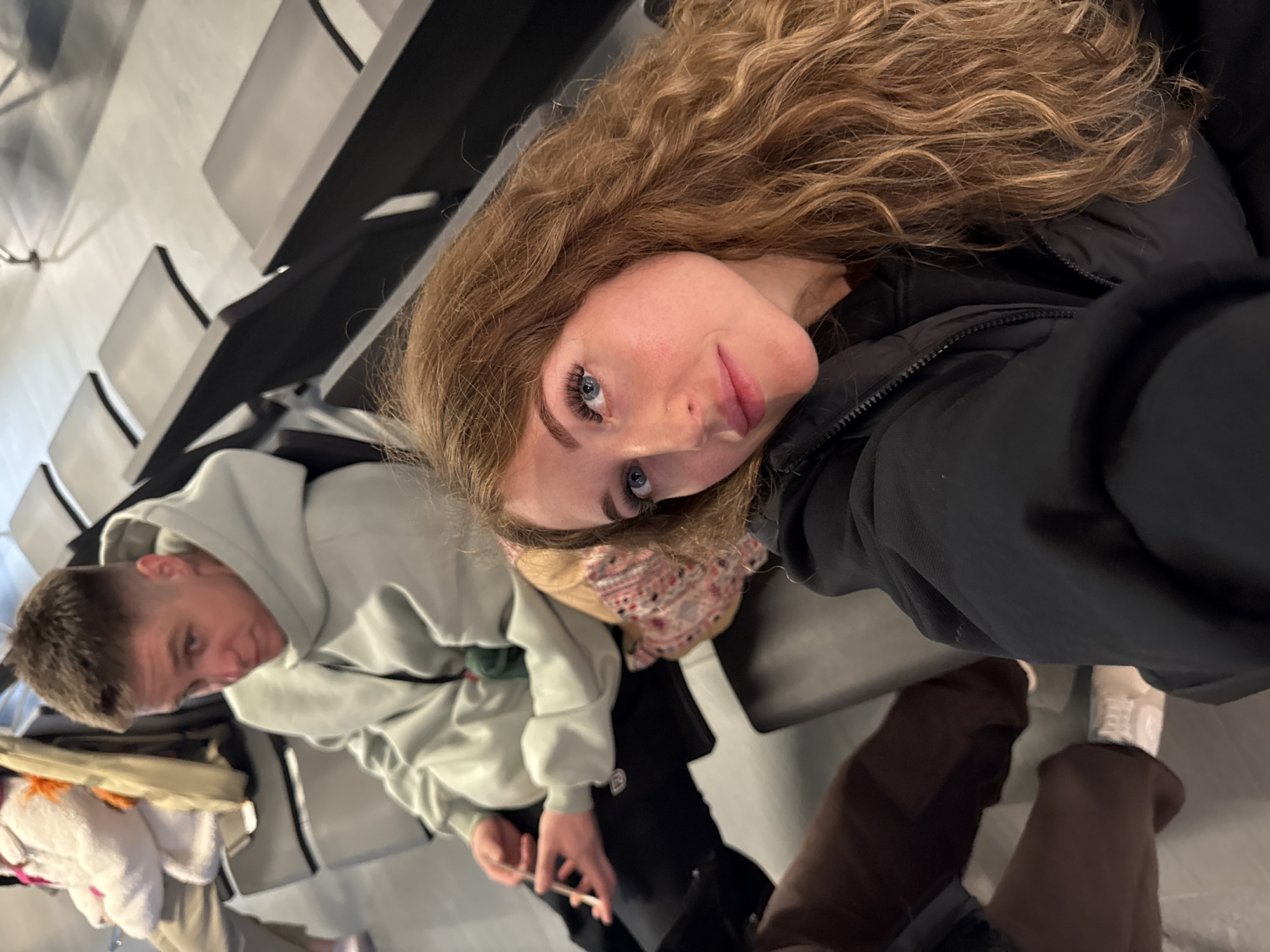 The image appears to be a selfie taken inside an airport waiting area. A young woman with long, curly blonde hair and blue eyes is facing the camera, wearing a black jacket. To her left is a young man wearing a light green hoodie. Both are seated on black airport-style seating. In the background are rows of empty chairs. The location is not Boldino, Russia.