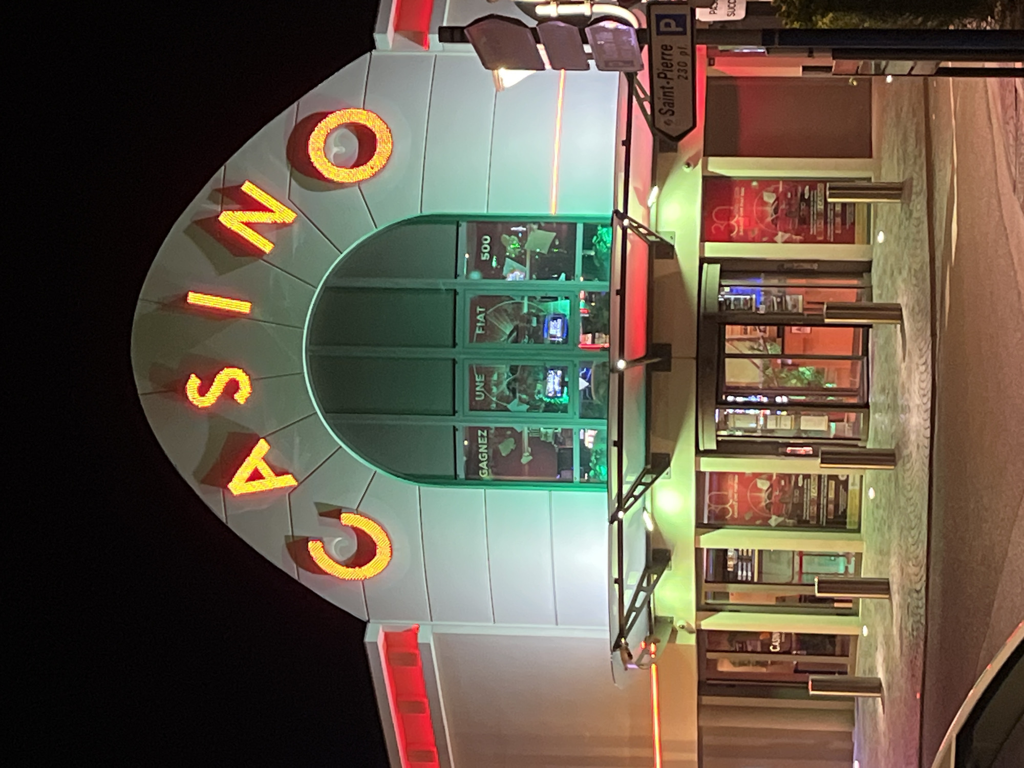 This is an exterior shot of the Casino Croisette in Cavalaire-sur-Mer, France, captured at night. The word "CASINO" is displayed vertically on the front of the building in large, illuminated orange letters. The building's design is modern, with a curved façade and large windows. The windows display slogans and advertisements, including references to winning a Fiat 500 and the tagline "GAGNEZ UNE FIAT."
A sign pointing to "Saint-Pierre" with a parking symbol is visible, indicating that the location is 230 meters from the Saint-Pierre area. The lighting is predominantly green and red, contributing to the atmosphere of a casino. A few people are barely visible inside of the building.