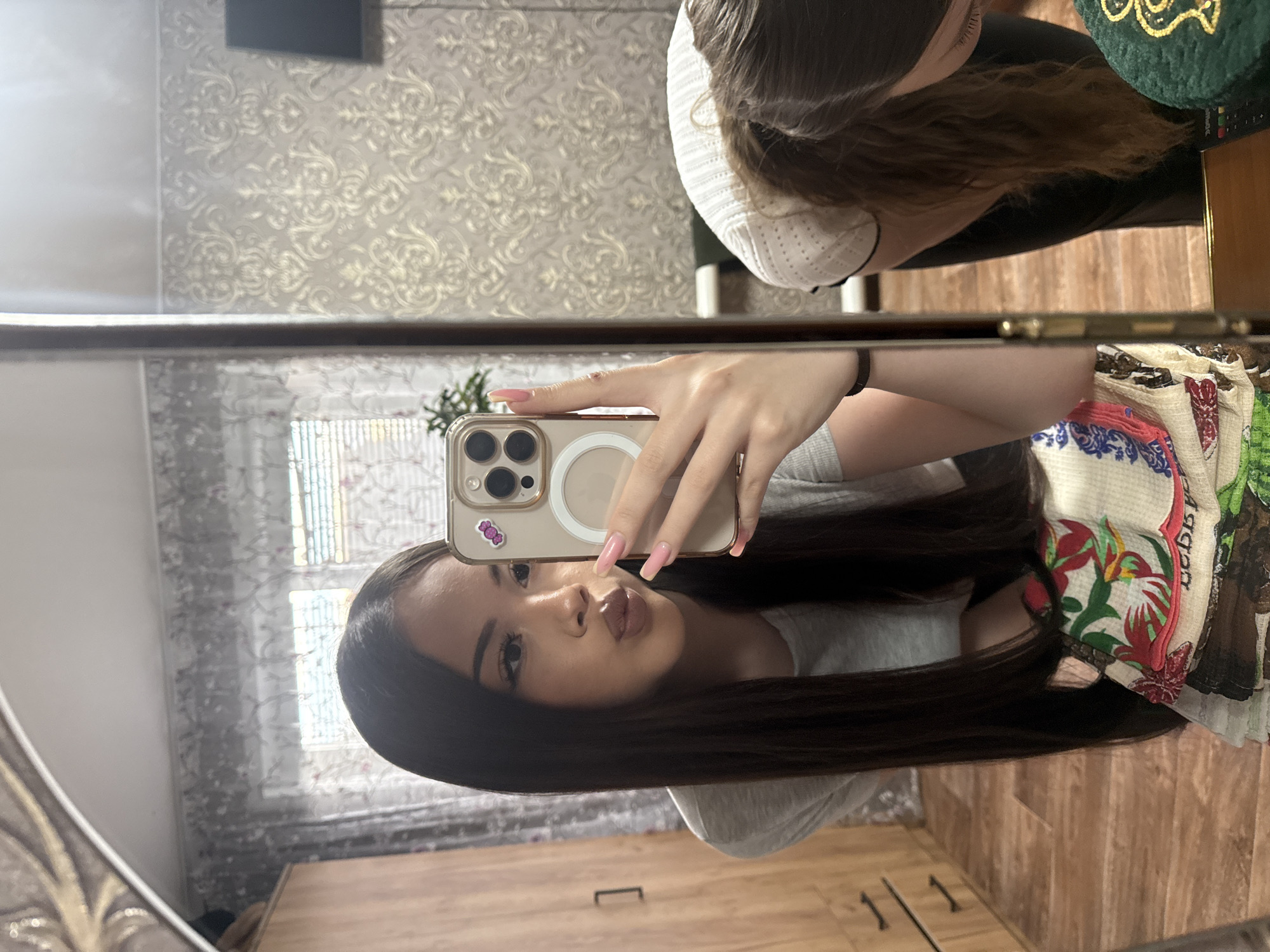 A young woman is taking a selfie in a mirror. She holds a smartphone in her right hand, with light pink polished nails. Her dark hair is long and straight. She is wearing a light grey t-shirt. The background includes a wall with patterned wallpaper and a dresser. The mirror reflects another person wearing a white hat and a table with some cloth. It is not possible to determine if the location is in the city of Konovalovka, Russia.