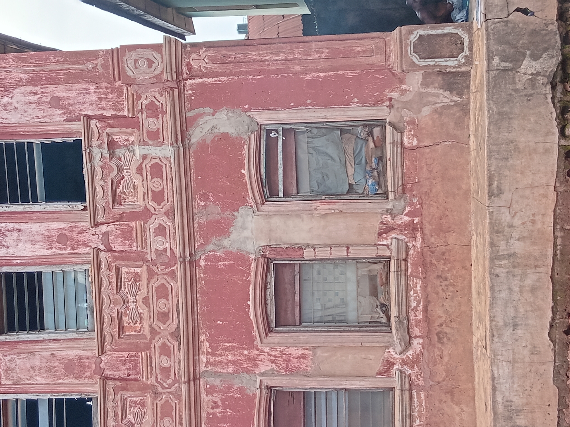 A multi-story building in Shagamu, Nigeria, displays weathered, reddish-pink painted facades with decorative, molded window frames. The building's structural integrity is compromised by cracking and crumbling. Windows contain opaque or partially obscured coverings. A vertical support structure along the right side shows substantial damage. A person is visible at the top of the frame.