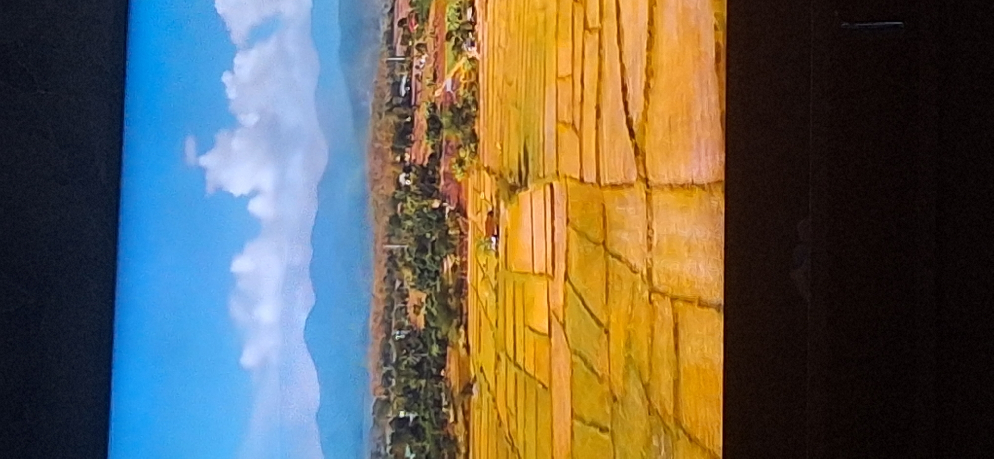The image shows an aerial view of a rural landscape with a bright blue sky and fluffy white clouds. In the foreground, there are geometric patterns of golden-yellow fields, likely rice paddies, divided by darker lines. In the midground, there is a lush green area with trees and scattered buildings, suggesting a village or small town. Further in the background, a range of blue mountains stretches across the horizon. The overall scene depicts a tranquil and cultivated agricultural area with a natural backdrop. This image does not show the city of Bila Tserkva, Ukraine.