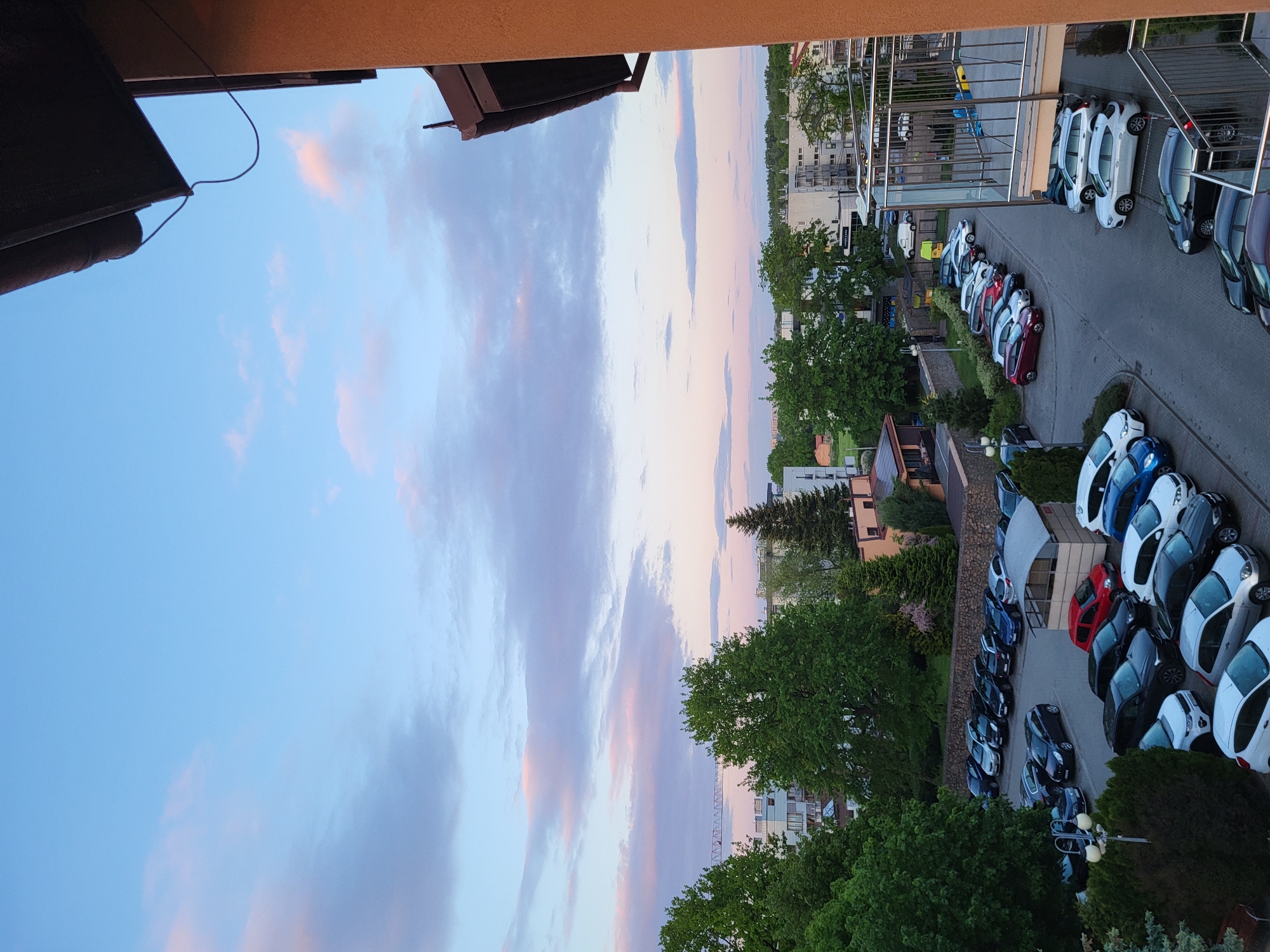 The image depicts an aerial view of a street in an urban setting in Unknown, Unknown at dusk. The scene is characterized by rows of parked cars, predominantly white and dark colored, aligned on both sides of the street. Trees with green foliage are interspersed between the buildings and cars, adding a touch of nature to the urban environment. The sky is a blend of blue and pink hues, indicative of a sunset or twilight setting. The perspective is taken from an elevated position, possibly a balcony or higher floor of a building. There are buildings that can be identified in the background.