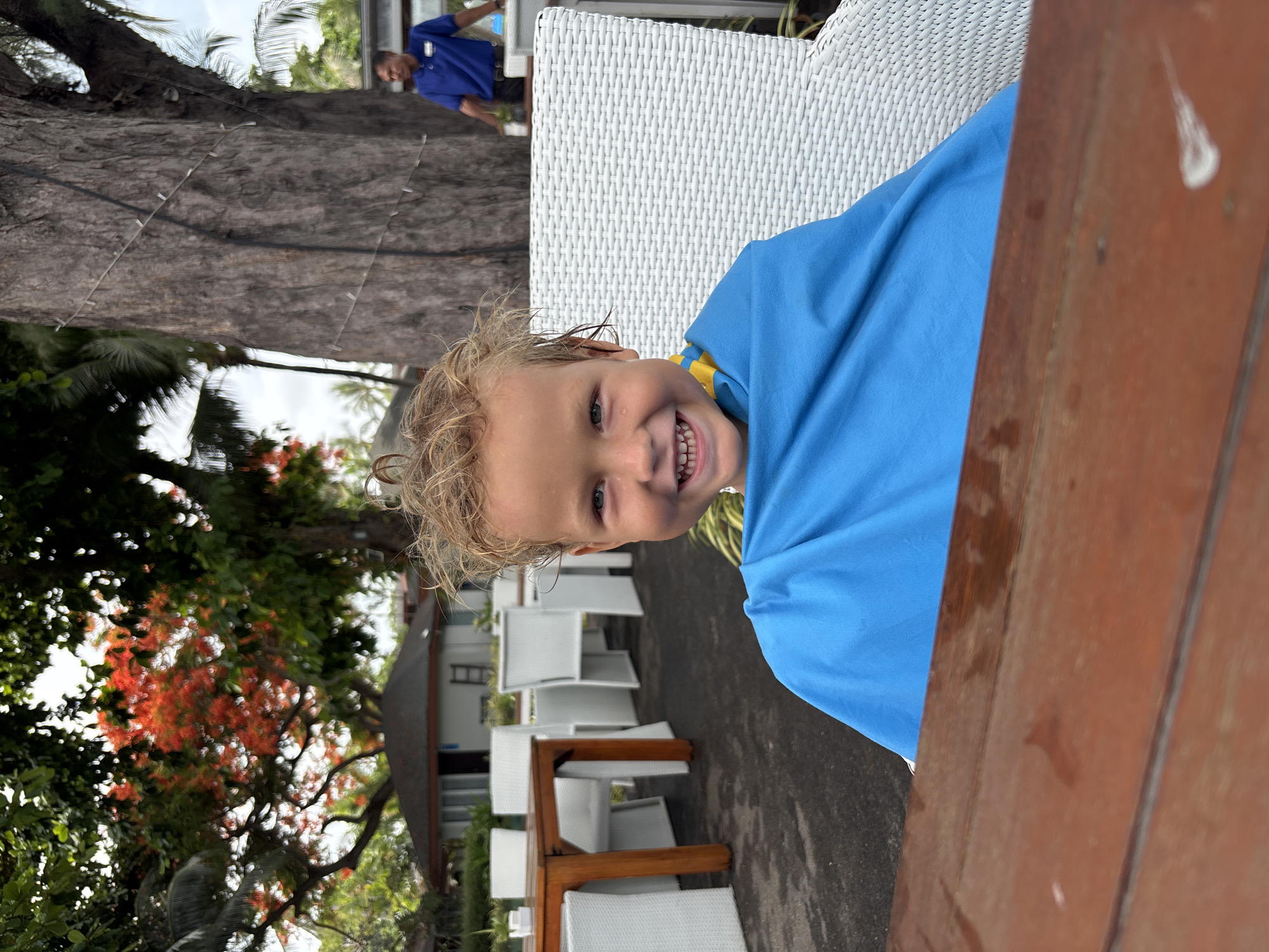 A young child wearing a blue towel is sitting at a table in an outdoor dining area at a location in Baan Chaweng, Thailand. A tree with red flowers is in the background, along with a building and other dining tables and chairs. A person in a blue shirt is partially visible in the background near a large tree. The photo appears to have been taken at an angle.