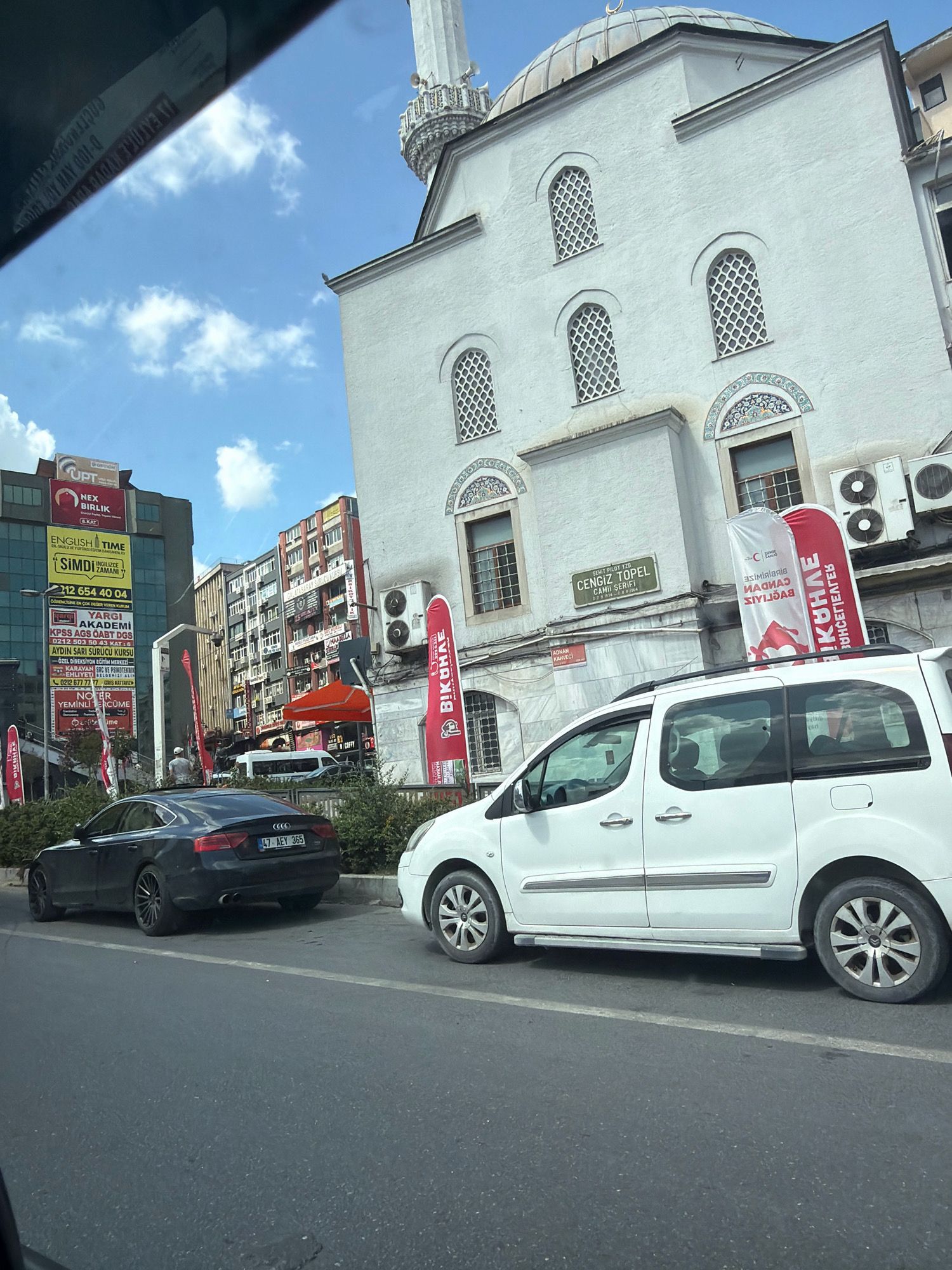 The image, taken from inside a vehicle, shows a street scene in Bahçelievler, Turkey. A white mosque with a domed roof and a minaret is prominent on the right side of the frame. In front of the mosque, two large vertical banners are displayed, one red and one white, with text in a foreign language. To the left of the mosque, two cars are parked on the side of the road: a black Audi sedan and a white minivan. In the background, multi-story buildings line the street, some with colorful facades and advertisements. The sky is partly cloudy.