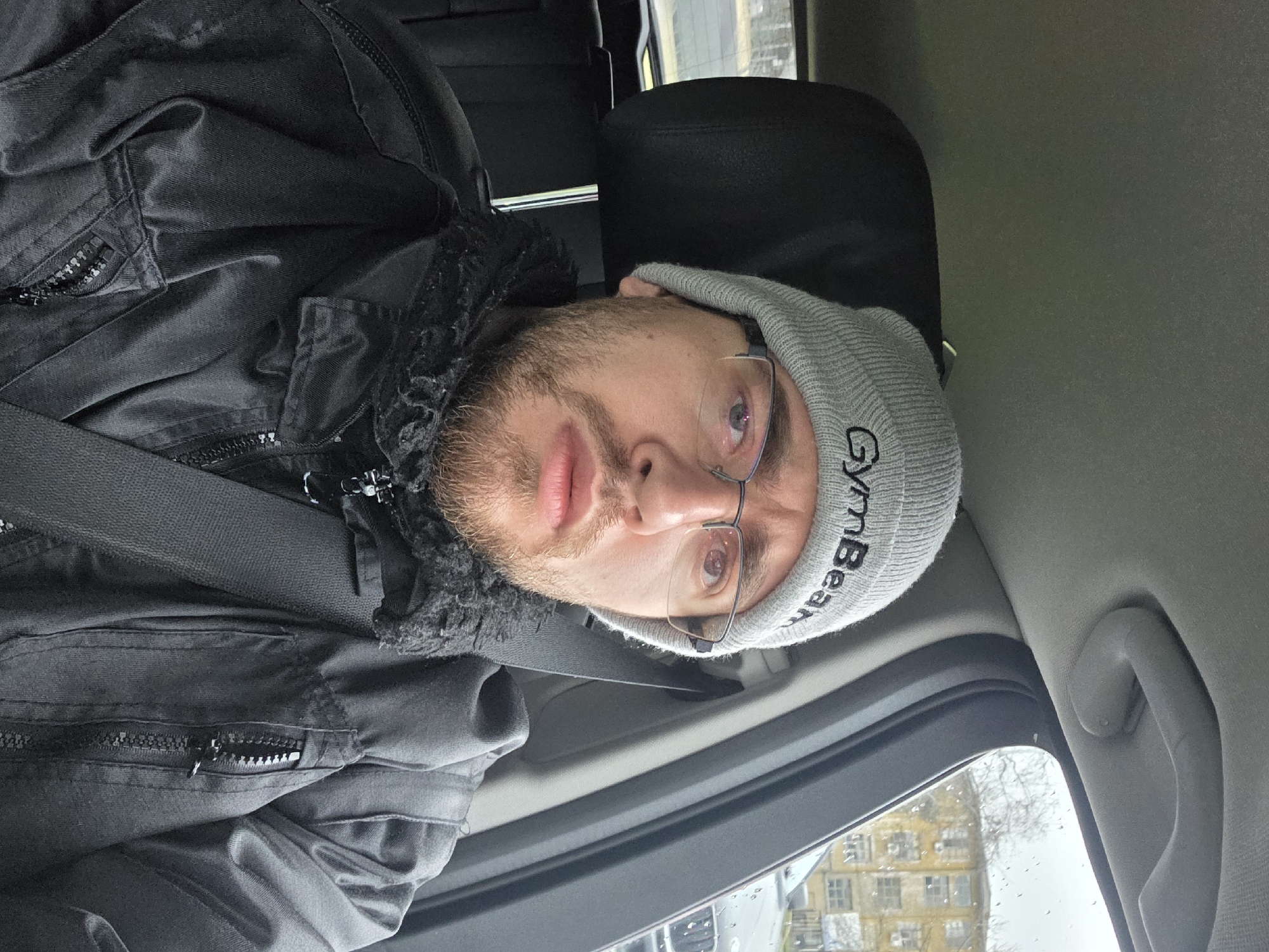 A man is in the driver’s seat of a vehicle. He is wearing a black coat, a gray beanie with the word “GymBeam” on it, and glasses. He is wearing a seatbelt. Outside the car window is a building. The building is in Plovdiv, Bulgaria.