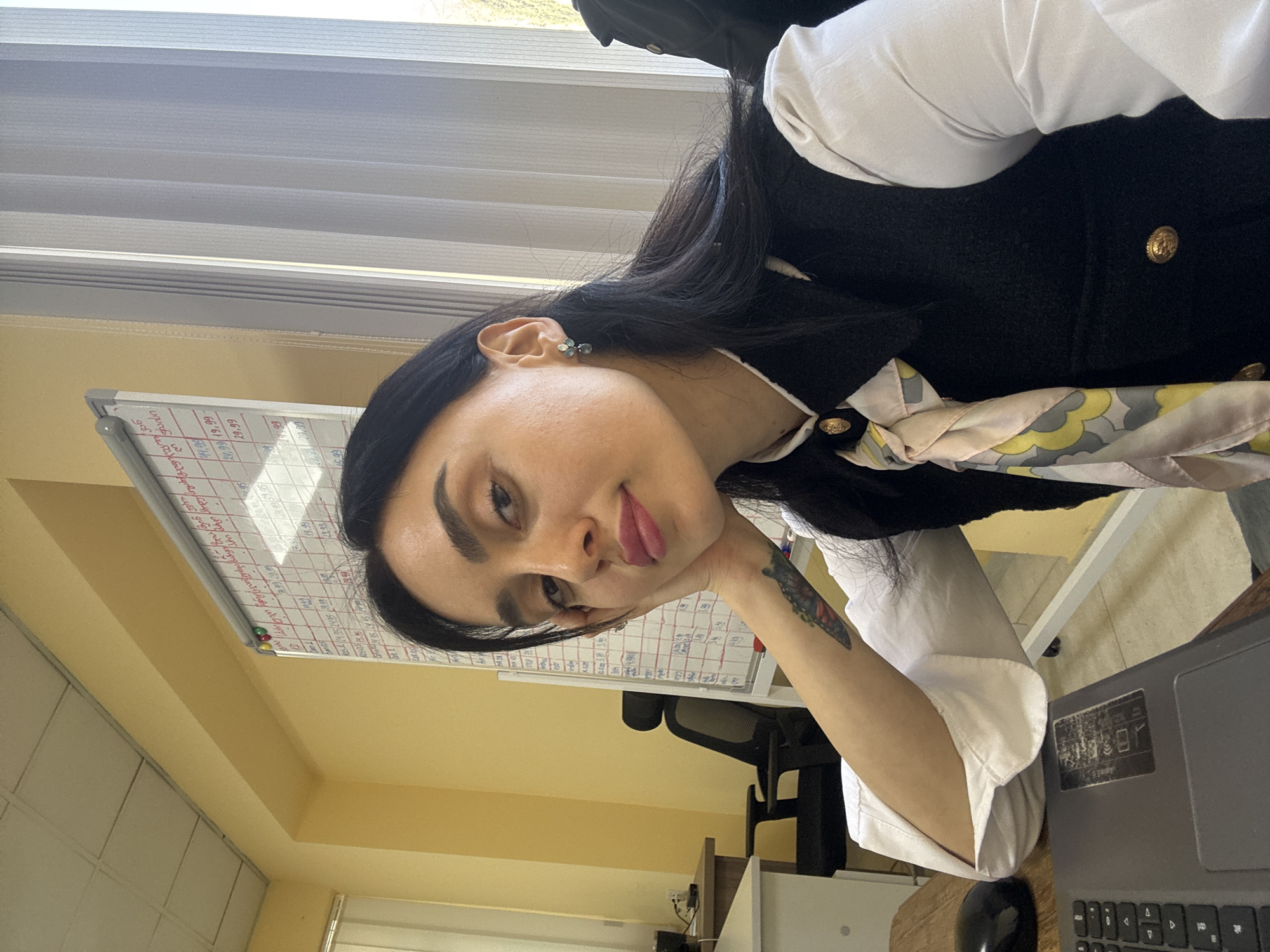 The image, correctly oriented, depicts a young woman seated at a desk in an indoor setting, likely an office or classroom in Tbilisi, Georgia, during the daytime. She is the central subject, resting her head on her right hand and looking towards the camera with a relaxed, subtle smile, her tongue slightly visible.
She has long, dark hair and is dressed in a white long-sleeved shirt under a black sleeveless vest with prominent gold buttons. A light-colored patterned scarf, featuring yellow, grey, and pinkish abstract shapes, is tied around her neck. She wears a small, possibly greenish-blue, stone earring on her right ear, and a colorful tattoo resembling a butterfly is visible on her left forearm.
The background includes a white dry-erase board covered with handwritten notes, numbers like "29.99", "19.99", "8.99", "60.99", and "55.55", and Georgian script, notably "დღე 1" and "დღე 2" (meaning "Day 1" and "Day 2"). Some entries on the board are crossed out. A black office chair is visible behind the board. The room features light yellow walls and a white ceiling, with a window fitted with horizontal blinds providing natural light from the upper left. On the wooden desk to her right, a grey laptop with an illegible sticker and a black computer mouse are present. Her expression conveys a sense of ease or thoughtful pause.