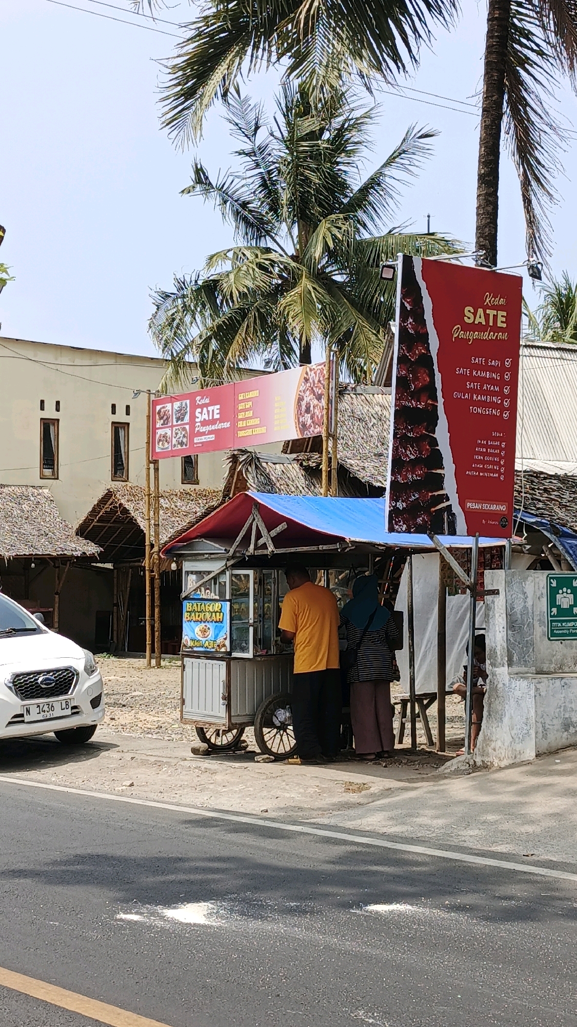 In the city of Desa Wonoharjo, Indonesia, an outdoor food stall is set up along a road. The stall, a cart with wheels, is covered by a blue and red tarp. A white car with license plate N 3436 LB is parked to the left of the stall. Two people are standing at the stall, one wearing an orange shirt and black pants, and the other a hijab and striped shirt with striped pants. A large red banner hangs above the stall, advertising various sate dishes. To the right of the stall, a green sign with a white icon of people is visible. The environment is sunny with palm trees visible in the background. The road in the foreground is asphalt.
