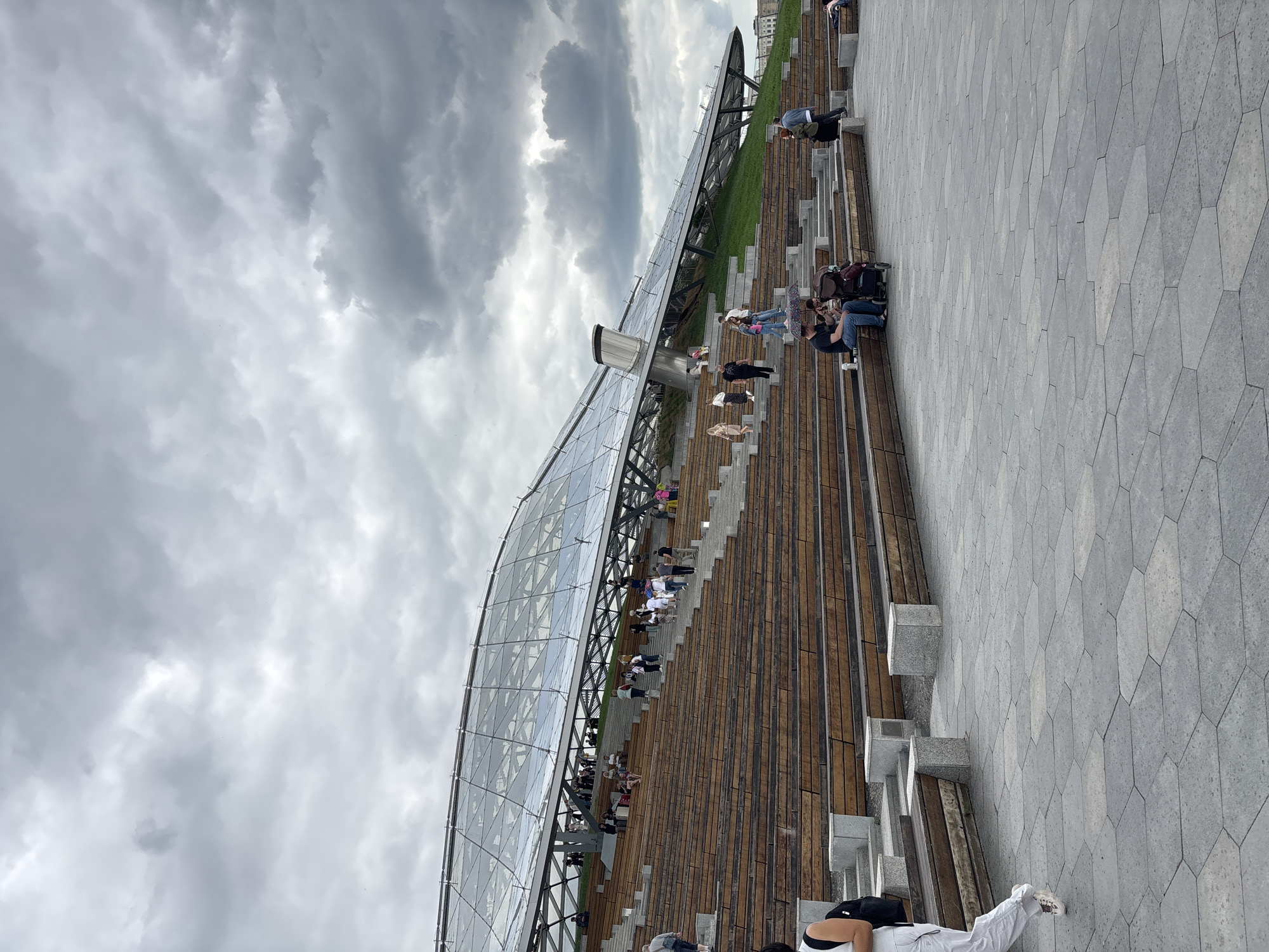 The image shows a sloping structure in Zaryadye Park in Moscow, Russia. The structure, a covered pedestrian bridge or viewing platform, features a glass roof supported by a metal frame. Many people are sitting and standing on the wooden steps that lead up the slope. Some individuals are interacting in small groups, while others appear to be relaxing or observing the surroundings. The sky is overcast with grey clouds. A paved area with a geometric pattern borders the wooden structure.