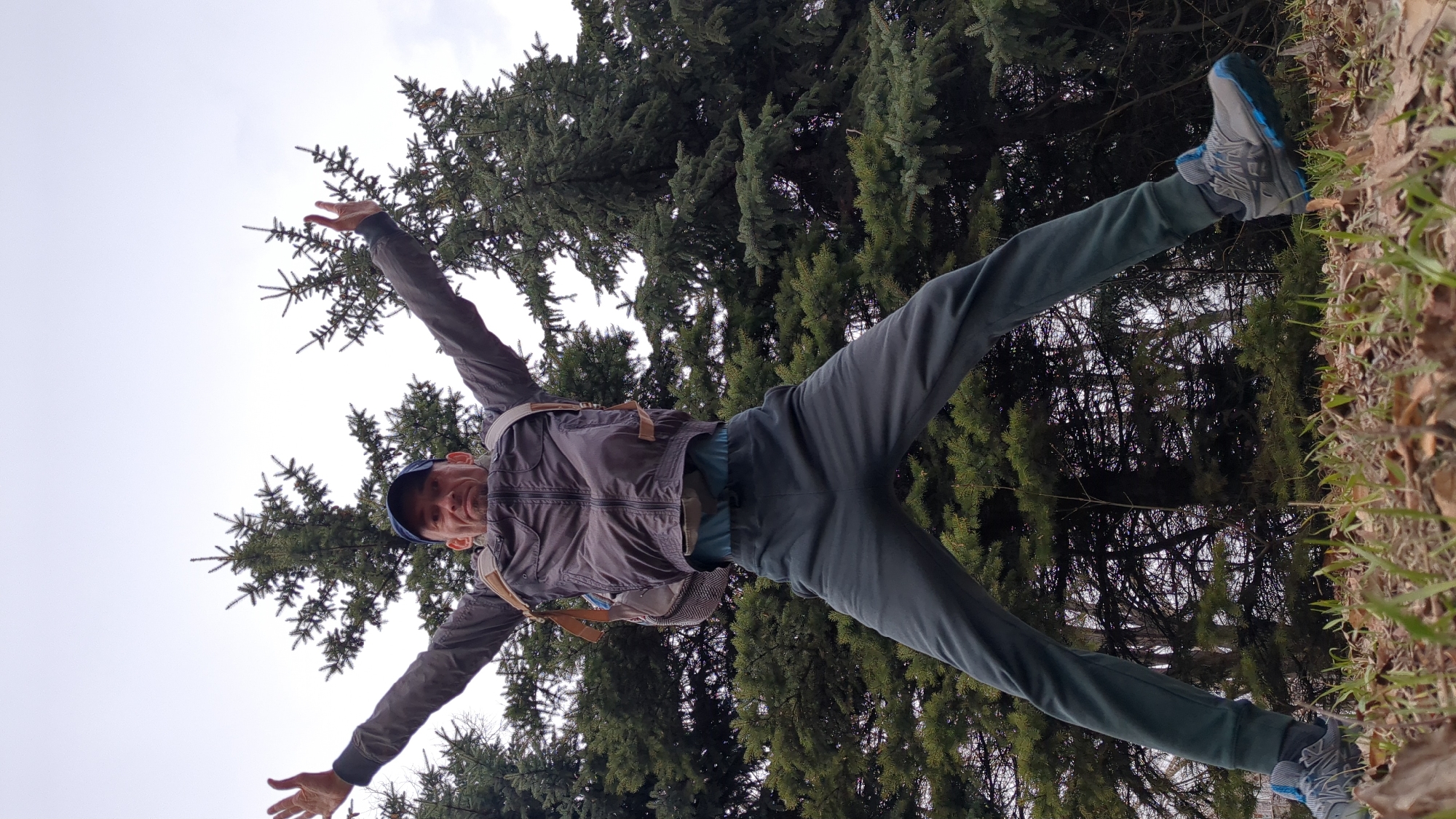A man stands with arms spread and legs apart, appearing to embrace the scene. He is wearing a dark jacket, sweatpants and blue-gray running shoes. A backpack is slung over his shoulders. A tree and sky are in the background and the ground to the right of the frame. The location is outdoors in Ulyanovsk, Russia.