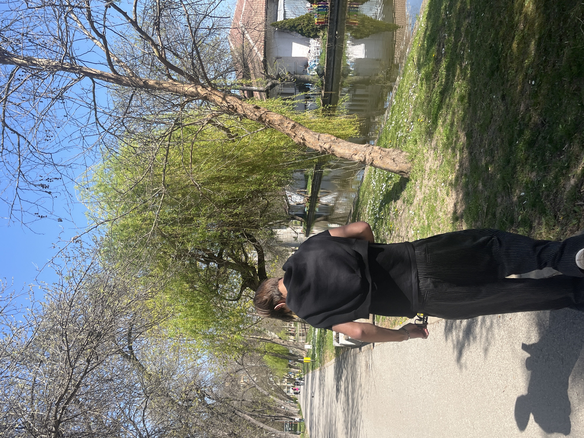A medium shot captures a person in black walking on a paved path lined with trees in Montana, Bulgaria. To the right of the path is a grassy area alongside a body of water, possibly a canal or small river, reflecting nearby buildings. The person is wearing a black top and vertically striped black pants, moving from right to left. The trees along the path are a mix of bare branches and new green foliage, suggesting early spring. The sky is blue. The overall impression is a peaceful scene in a city park.