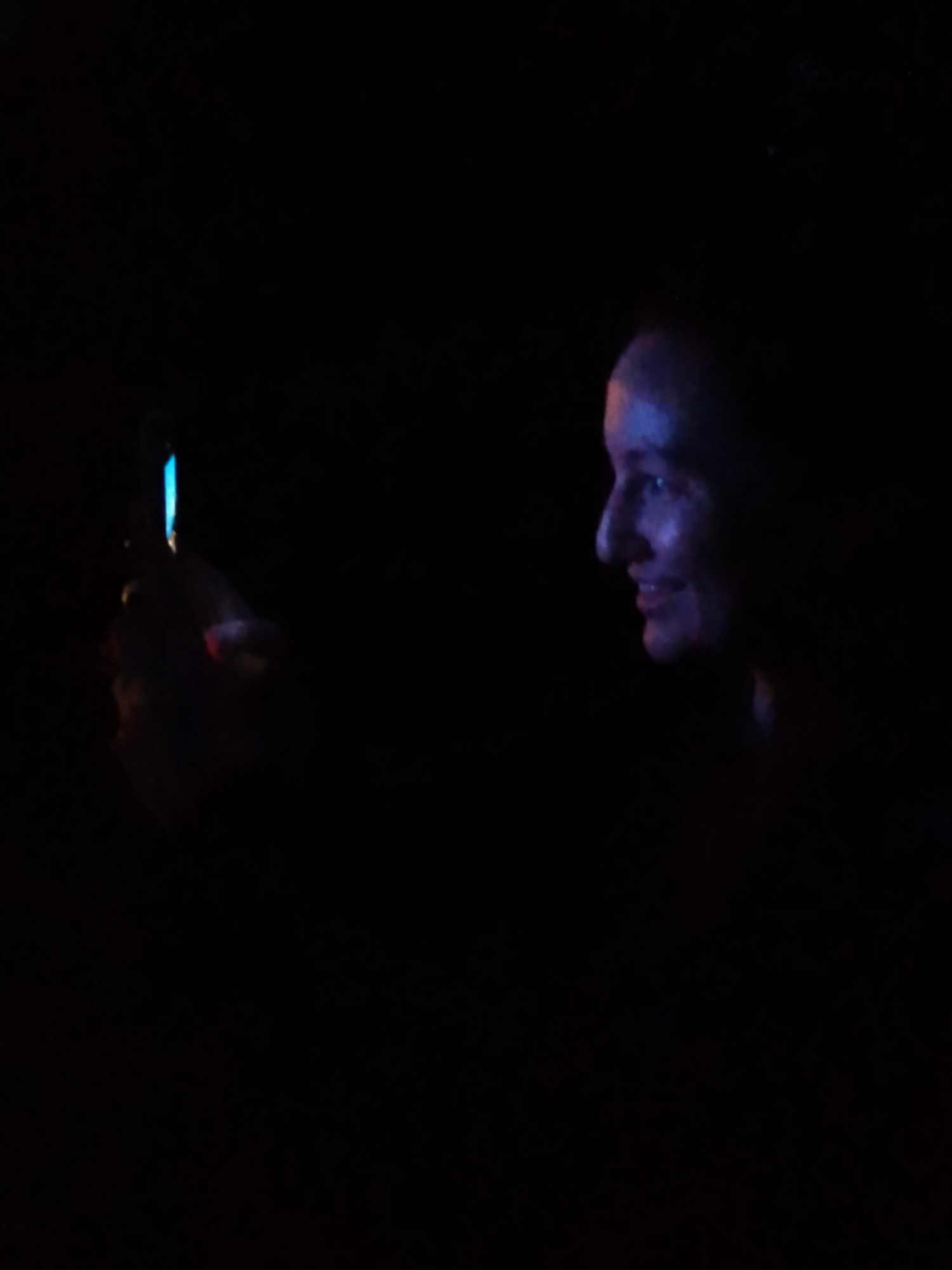 In a dark environment, a person's profile is visible on the right side of the image, illuminated by blue and purple light. Their face is turned to the left, with their mouth slightly open in a smile. On the left side, a hand holds a small object that emits a bright blue light. The background is completely black, suggesting the scene is set at night or in a dimly lit indoor space. The image does not provide any specific location in Schwerin, Germany, or any details that would suggest it.