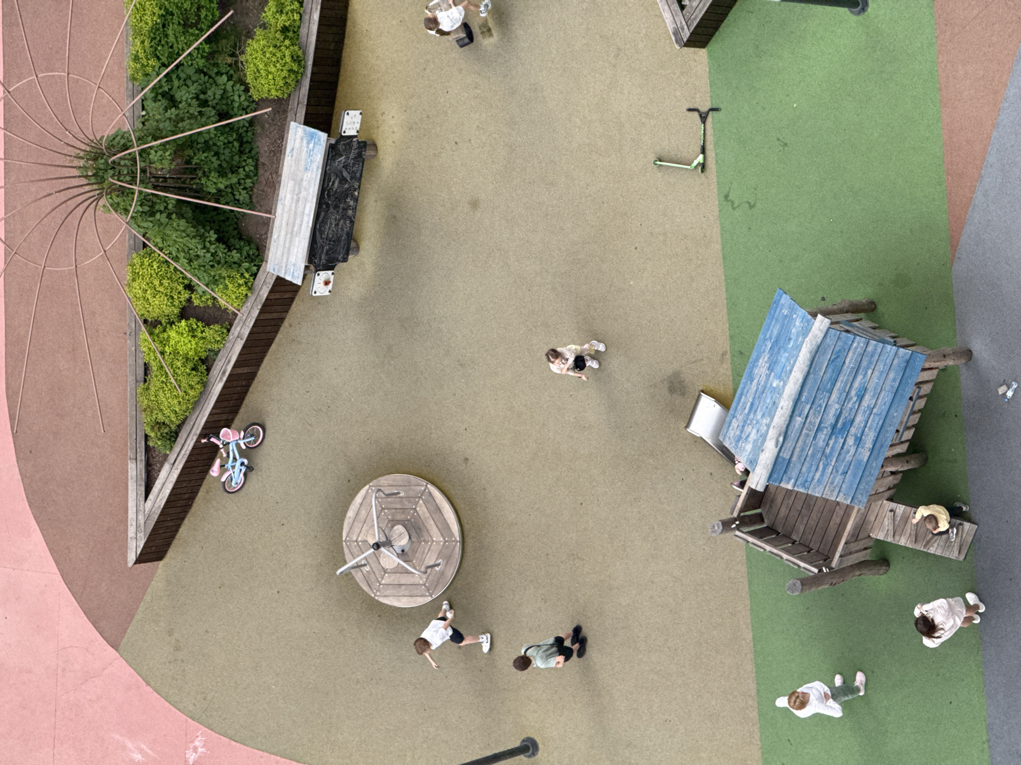 Aerial view of a playground in Perm, Russia. Children are present in the playground with various play structures, including a spinning platform, wooden playhouse with a slide, and planted areas with vegetation and an ornamental structure. There's a bicycle, scooters, and scattered objects on the ground. The ground is covered with different colored surfaces.