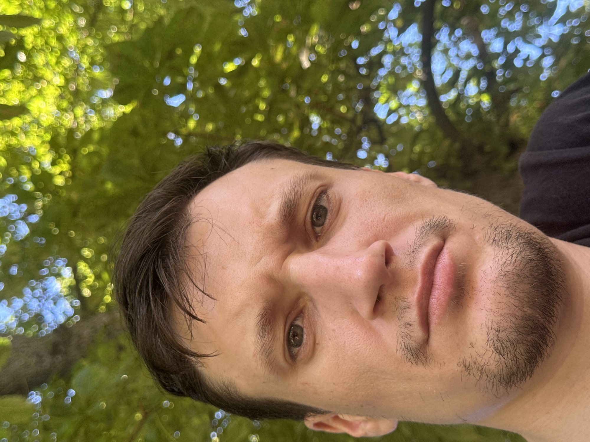 The image is a close-up selfie of a man with brown hair and a short beard, taken outdoors. He has a slightly furrowed brow and is looking at the camera. The background is out of focus, showing lush green foliage with bokeh effects from sunlight filtering through the leaves. The man is wearing a black t-shirt. There is no indication of the city of Makiv, Ukraine, in the image.