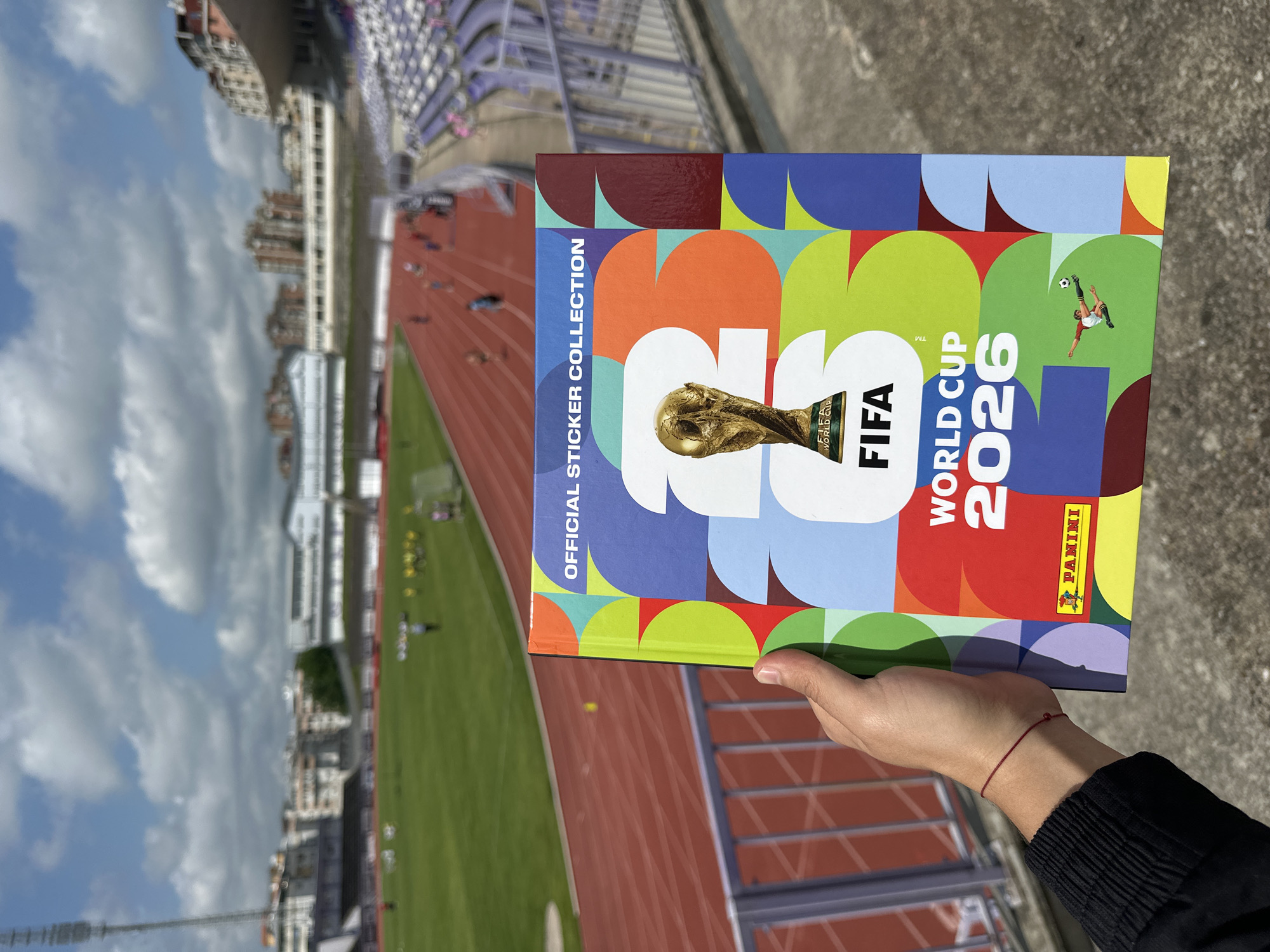 A person's hand, adorned with a simple red string bracelet, holds a colorful FIFA World Cup 2026 Official Sticker Collection album. The album's cover features a vibrant, abstract design with geometric shapes in various colors, prominently displaying the FIFA World Cup trophy, the FIFA logo, and the text "WORLD CUP 2026." "OFFICIAL STICKER COLLECTION" is written vertically on the left side, and the Panini logo is visible at the bottom right. A small illustration of a soccer player is also part of the design.
The scene is set outdoors at a sports stadium in Veliko Tarnovo, Bulgaria, under a bright sky with scattered clouds, indicating daytime. In the background, a red running track stretches around a green field where several blurry figures, possibly athletes or individuals, are visible. Stadium seating, predominantly purple and white, can be seen to the right. Beyond the stadium, residential buildings with distinct architectural styles rise against the horizon. The image offers a perspective from a higher vantage point within the stadium.
