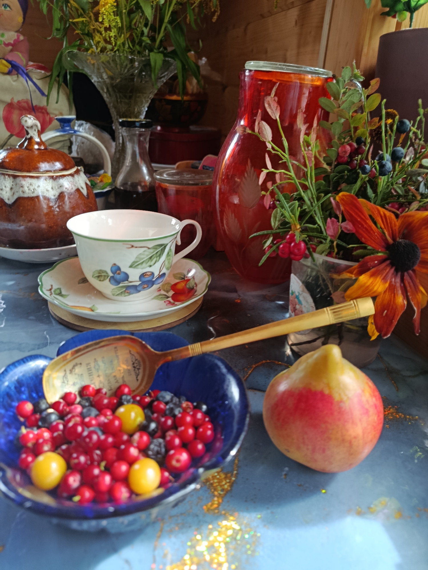 A still life arrangement is captured from a slightly elevated, medium shot perspective, presenting an eclectic collection of objects on a textured, blue-gray surface speckled with gold glitter. The scene is illuminated with warm, natural light, casting soft shadows.
Dominating the center-left is a rustic, brown ceramic sugar bowl with a white, textured rim and a decorative lid featuring a white knob. It sits on a white plate. Adjacent to it, a delicate porcelain teacup and saucer, adorned with a blue blueberry motif and a green rim, is placed on a thin, brown circular coaster.
To the right of the teacup, a large, translucent red glass pitcher, intricately etched with a floral design, is filled with a clear, red liquid. The pitcher is adorned with a bouquet of greenery and berries, including deep blue blueberries and small, red berries, alongside a vibrant orange-black-eyed Susan flower. The bouquet is partially contained within a clear plastic vase.
In the foreground, a deep blue, scalloped-edge bowl overflows with a colorful assortment of small berries, primarily bright red cranberries, interspersed with dark blue blueberries and a few bright yellow berries. A golden, ornate spoon with an inscription is partially submerged in the berries, its handle extending diagonally across the bowl.
To the right of the berry bowl, a single, pear-shaped fruit, transitioning in color from yellow at the stem to red at the base, rests on the surface. In the background, hints of wooden paneling and a glimpse of a large, ornate glass vase holding more greenery can be seen. The overall impression is one of abundance and a rustic, autumnal aesthetic.