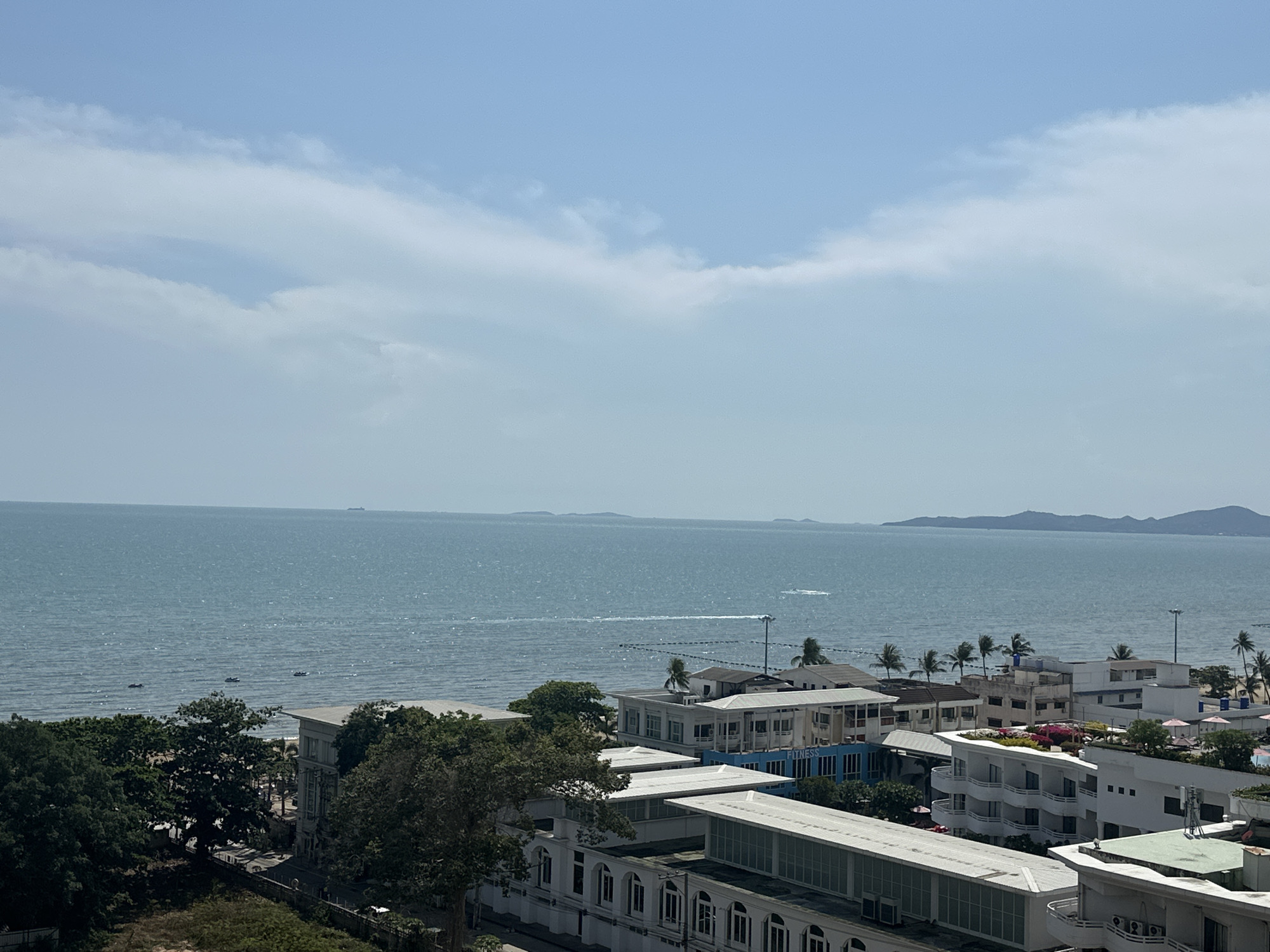 The image shows a view overlooking part of Pattaya City, Thailand. The foreground features buildings, mainly white, with some palm trees. The middle ground consists of the ocean, with visible waves and a few boats. In the background, there are islands on the horizon. The sky is light blue with some clouds. No people are directly visible.