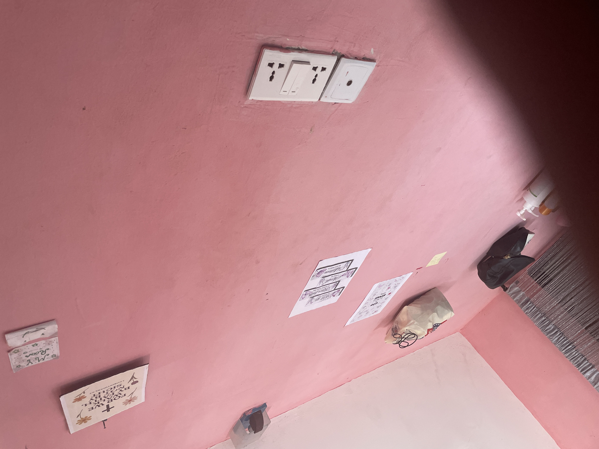The image displays a close-up, low-angle shot of a pink wall in what appears to be a residential interior, possibly in Iyafin, Nigeria, judging by the context provided. The wall is textured and shows some minor imperfections.
Mounted on the wall are several objects. To the upper left, there are two small pieces of paper with writing on them, possibly notes or decorations. Below these, a white paper with floral designs and text, including a cross symbol, is affixed to the wall. Further down and to the left, a clear plastic bag containing dark objects is attached to the wall.
In the center of the frame, there are two diamond-shaped prints with purple accents and text, suggesting decorative items or perhaps postcards. Below these, another piece of paper with writing and symbols is visible. A light-colored tote bag with black handles is hanging on the wall in the lower left quadrant, and near it, a black bag is placed on a surface.
On the right side of the wall, there is a white electrical outlet with two sockets and a switch above them, all mounted on a white plate. Below this, another white electrical fixture, possibly a socket or a dimmer switch, is installed.
In the lower portion of the image, a white pump bottle and another bottle are visible, suggesting toiletries or cleaning supplies. The foreground is partially obscured by a shimmering, curtain-like material, possibly a beaded curtain or tinsel. The lighting suggests an indoor environment, and the overall impression is that of a somewhat cluttered personal space.