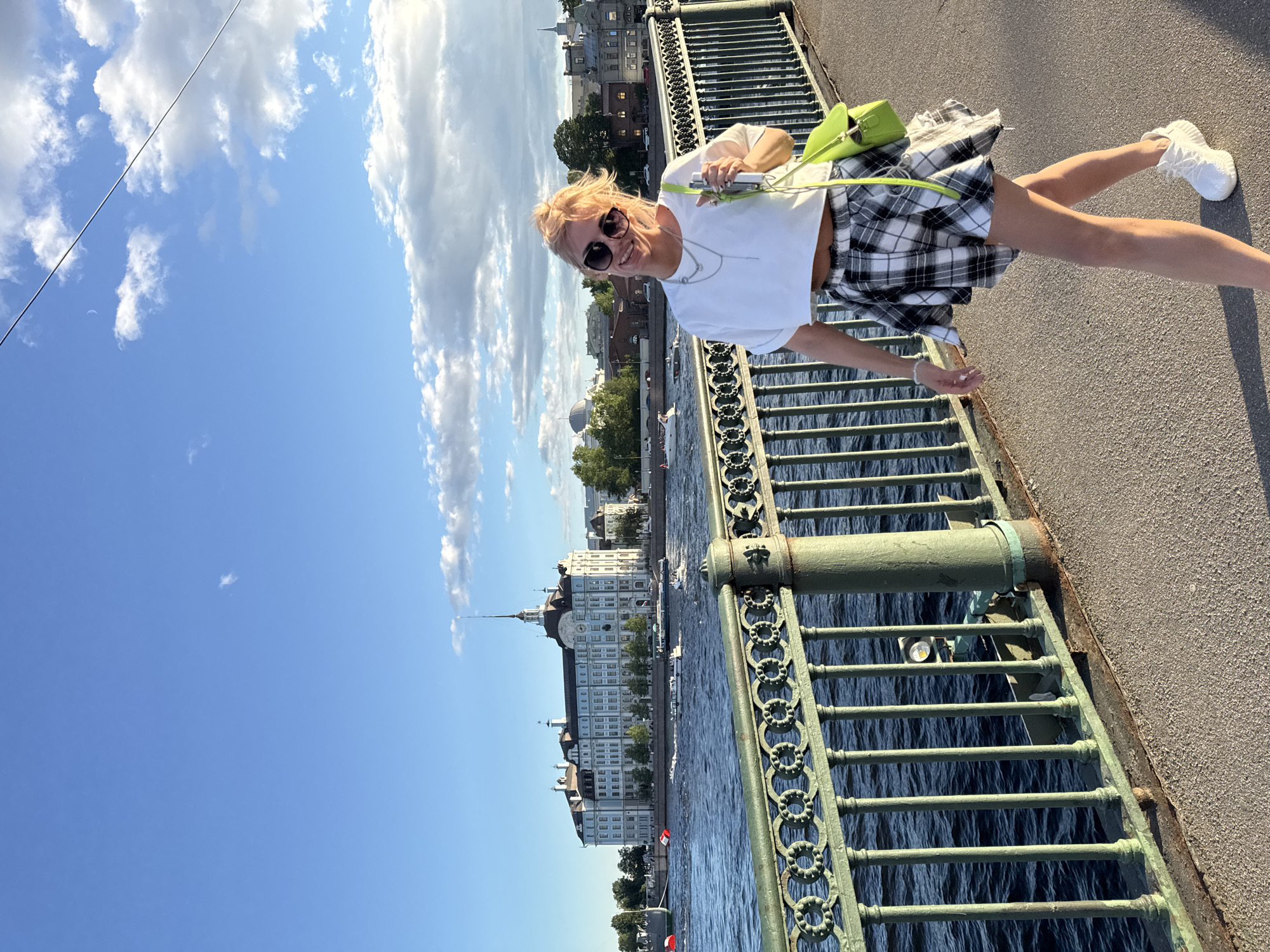 A blonde woman with sunglasses, a white crop top, and a plaid skirt walks along a bridge in front of a body of water. She carries a bright green purse. In the background, there is a large blue building with a spire and several boats on the water. The sky is blue with some clouds.