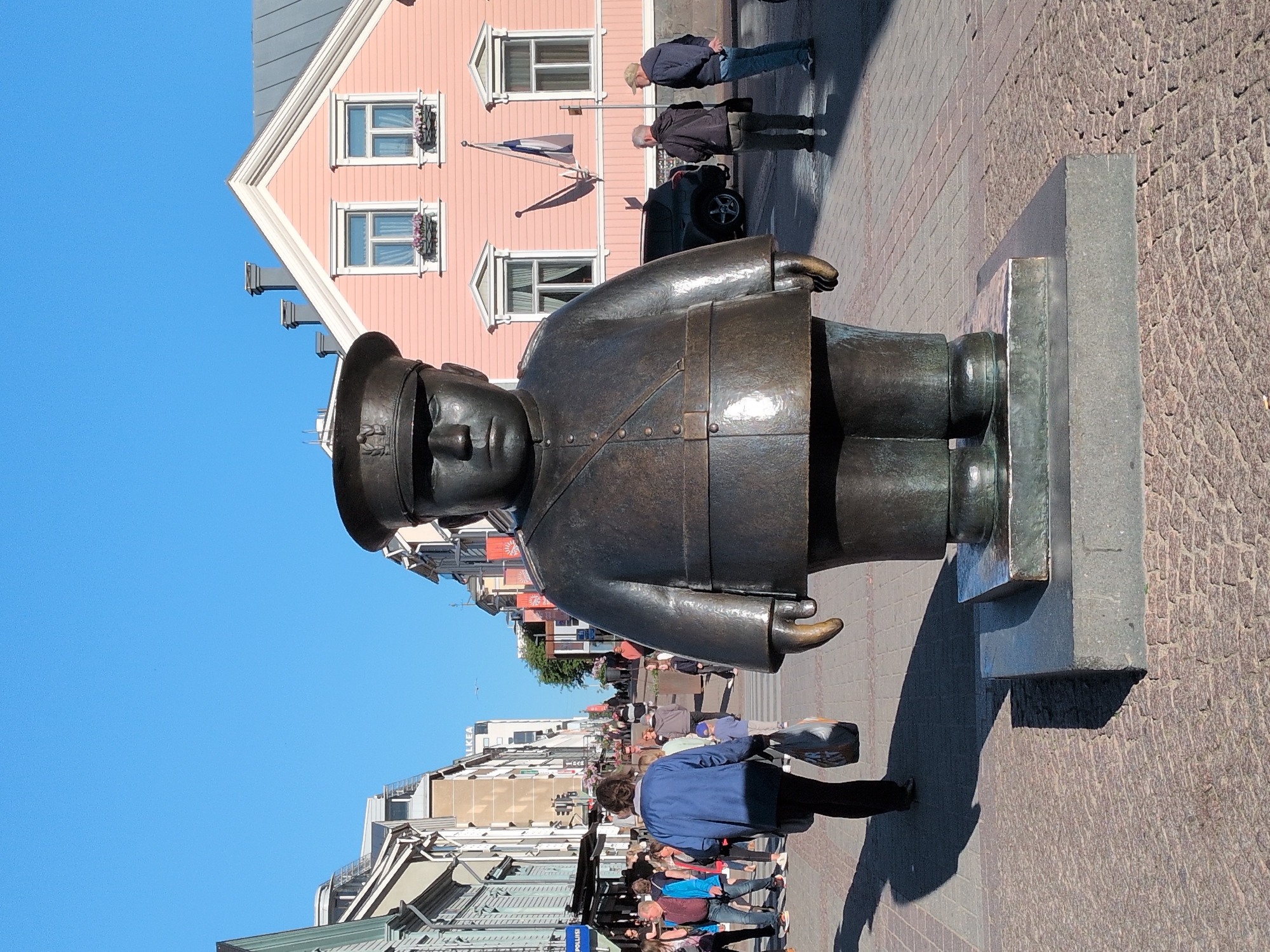 In Oulu, Finland, a large bronze statue of a stout man in a uniform with a hat stands prominently in the foreground of a pedestrian street. The statue faces forward and is positioned on a grey stone base. Behind the statue, a pale pink building with white trim and symmetrical windows is visible, adorned with flags. The street is paved with cobblestones, and several people are walking in the background. To the left of the statue, a woman in a blue jacket carries a shopping bag. A dark SUV is parked on the right side of the street. The sky above is clear and blue.