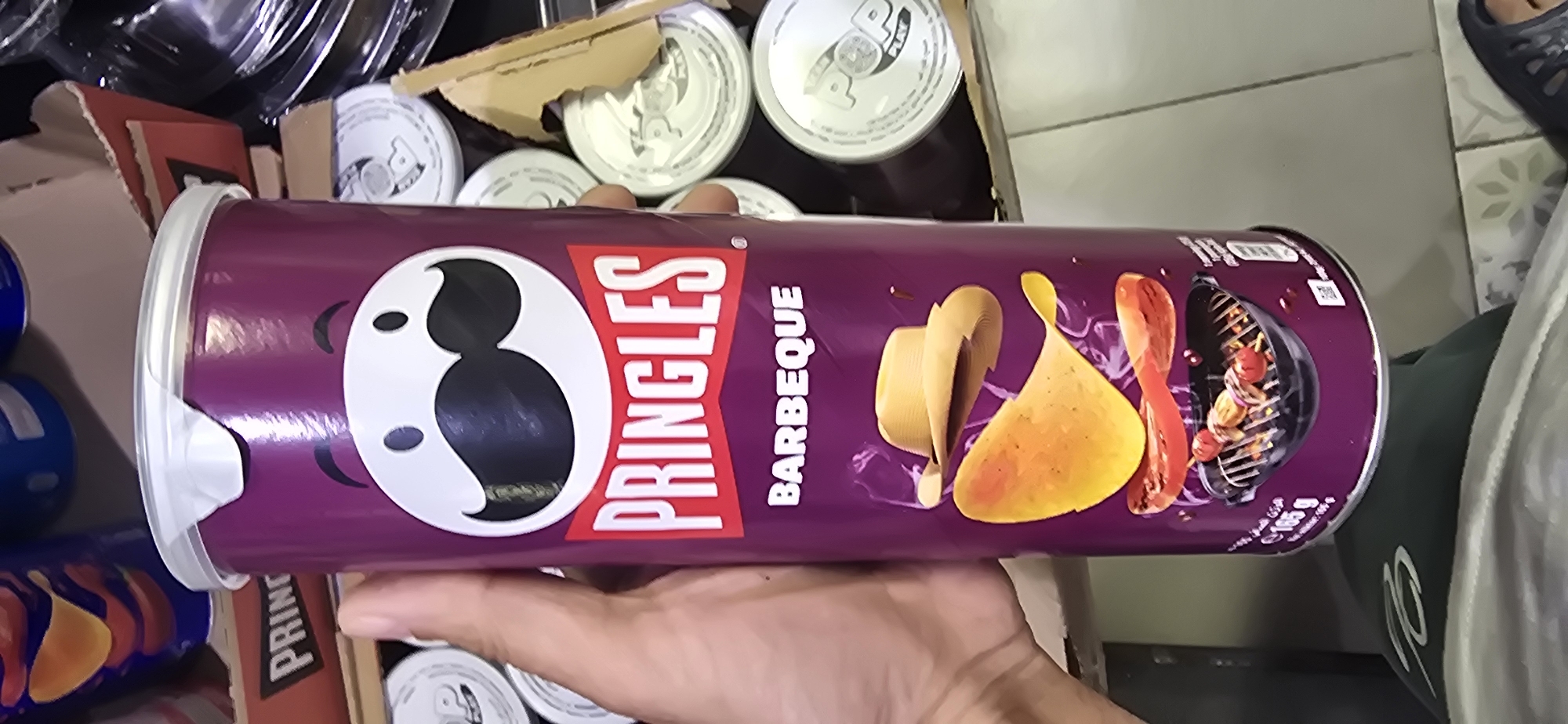 A person's hand holds a Pringles BBQ flavor potato chip can. The can is purple with the Pringles logo and an illustration of potato chips, hot dogs, and a grill. The background is a bit blurry, showing other Pringles cans, some plastic containers, and what appears to be a box. The setting seems to be indoors, possibly a pantry or storage area. The lighting is bright, suggesting daytime. There is text visible on the can: "PRINGLES" and "BARBEQUE." The overall impression is casual, possibly someone is showing off a snack they have.
