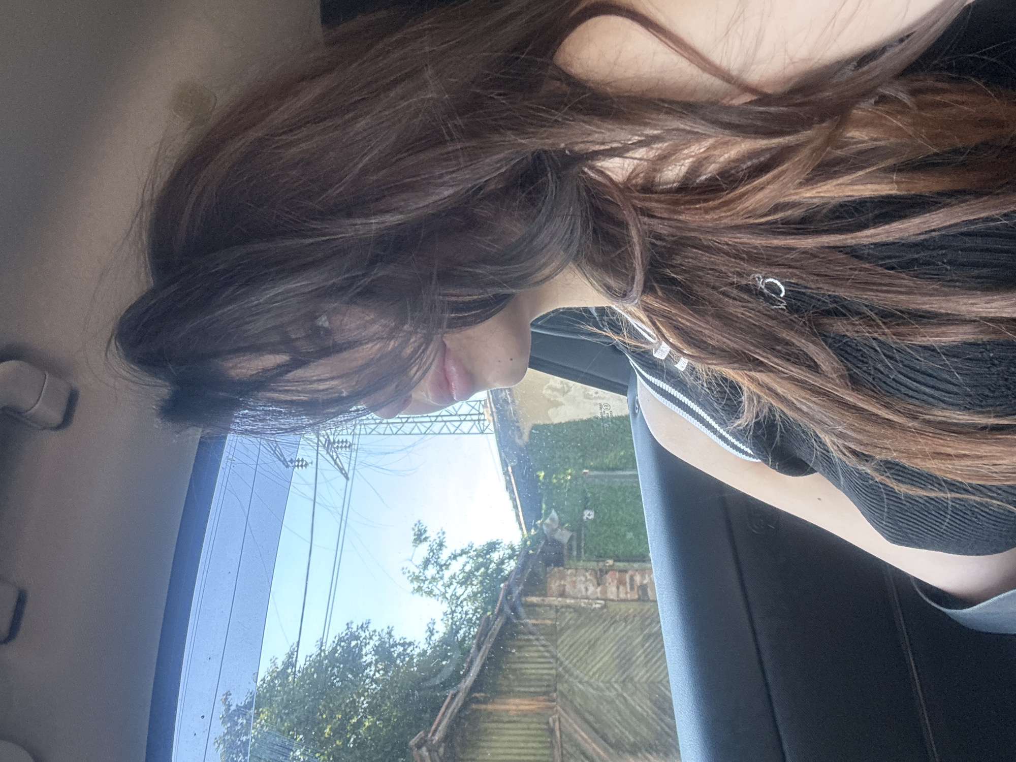 A woman is captured in a car, looking out the passenger window. Her long, dark brown hair is partially obscuring her face. The background through the window reveals a street scene in Sofia, Bulgaria, with a weathered wooden building and a power line tower. The interior of the car, with its black door panel and gray ceiling lining, is also visible. The woman is wearing a black tank top with white trim.