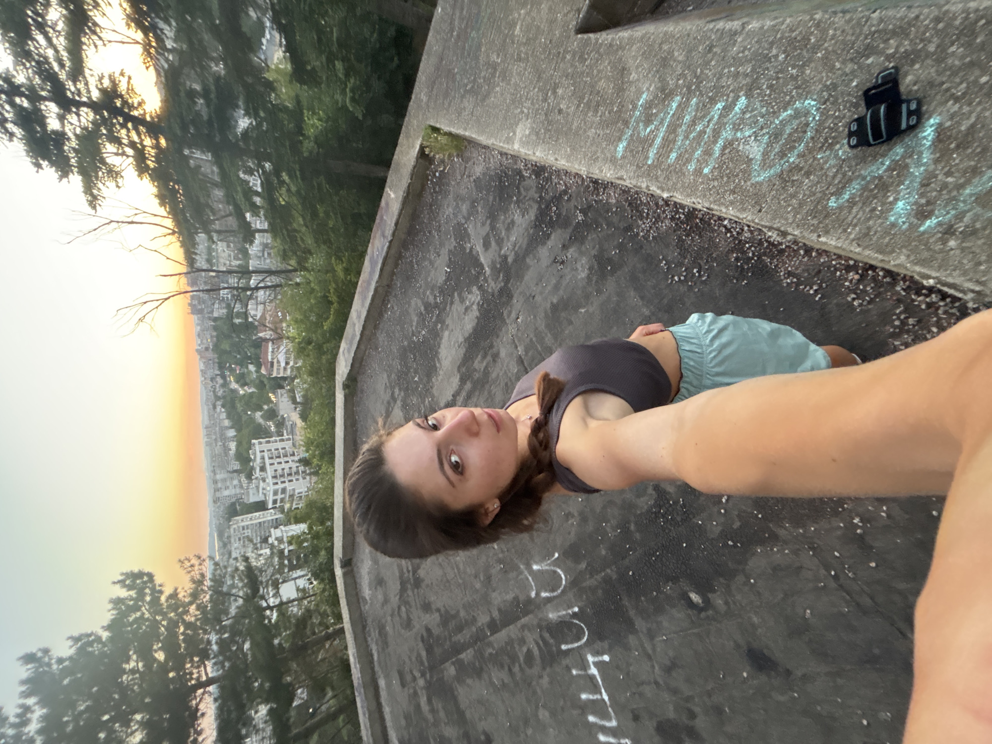 The image shows a young woman with brown hair in a braid, wearing a dark gray tank top and light blue shorts. She is holding her phone out, taking a selfie. She is standing on a flat, concrete surface with some debris and chalk markings. In the background, there is a panoramic view of Varna, Bulgaria, with buildings, trees, and a sunset in the sky. The location appears to be a high vantage point overlooking the city, possibly a viewpoint or an abandoned structure. There is a black object with straps lying on the concrete ledge in the lower right portion of the image.
