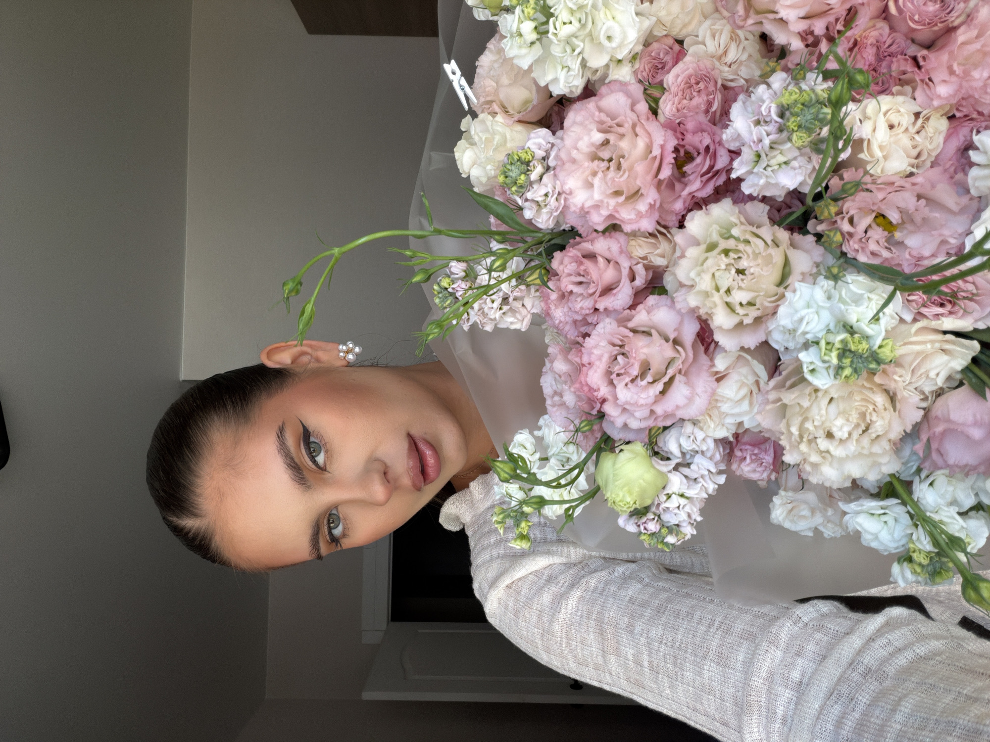The image shows a fair-skinned woman with brown hair pulled back, wearing an off-white textured sweater and pearl earrings. She has green eyes and distinct eyeliner. She is holding a large bouquet of pink and white flowers. The background is a plain grey wall. There is no evidence in the image to suggest this photograph was taken in Krasnoyarsk, Russia.