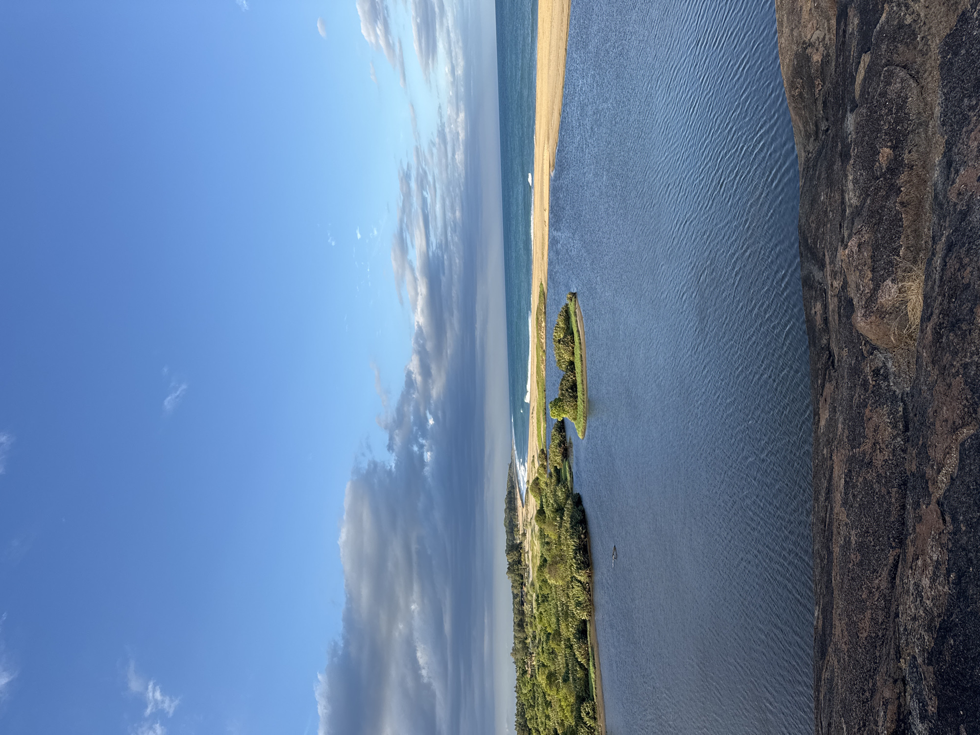 This is an aerial, landscape photograph of the coastline in Ulla, Sri Lanka. The scene is dominated by a body of water in the foreground, a strip of sandy beach, and the ocean beyond. Dense green foliage covers the land between the foreground water and the beach. The sky is blue with scattered clouds, some of which are dark and heavy, suggesting recent or impending rain. In the water, there is a small island covered in greenery. A bird is visible flying low over the water. The foreground consists of a rocky outcrop. The overall impression is one of a natural coastal environment.