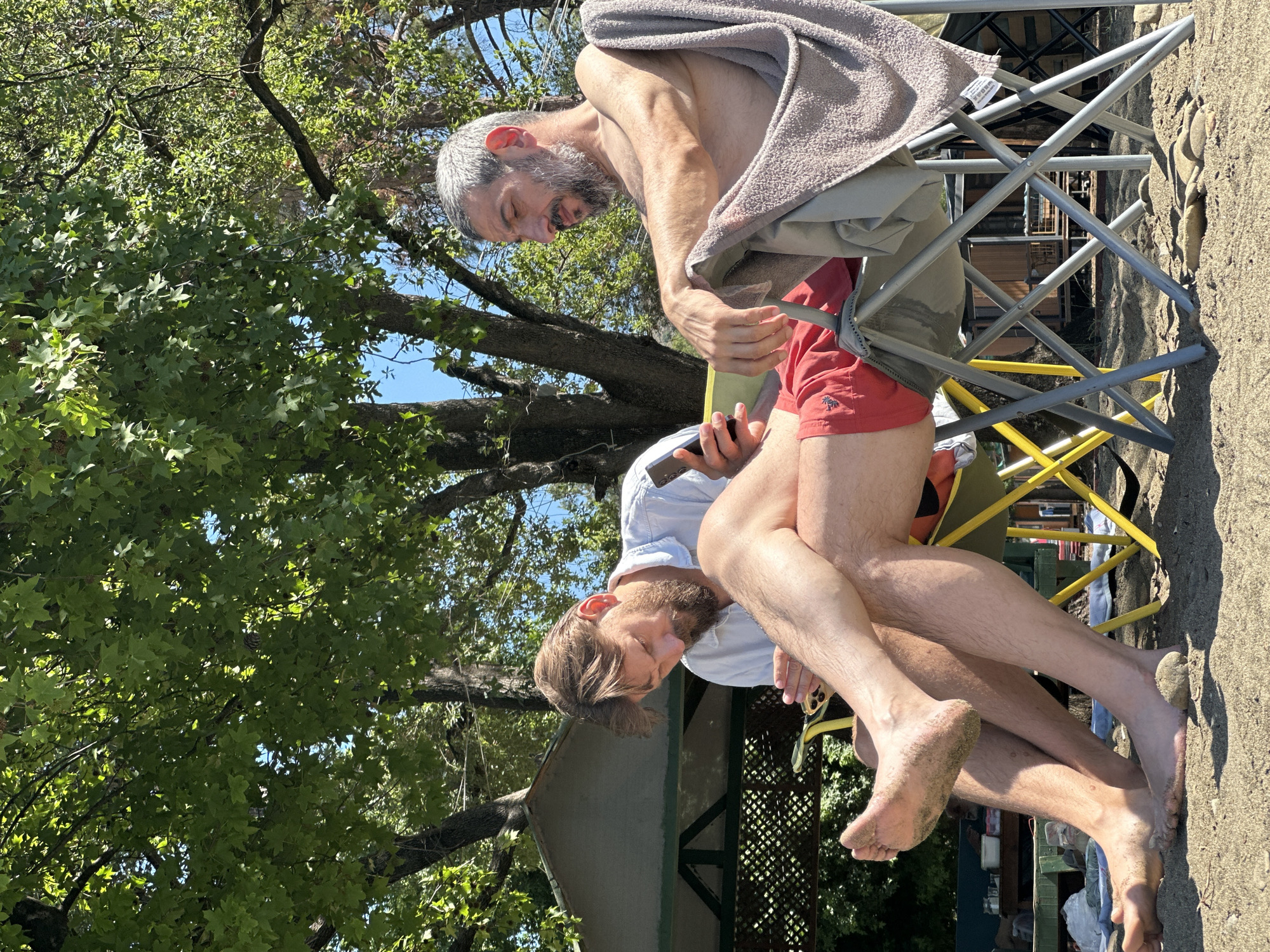 Two men sit side-by-side on foldable chairs outside under tree cover. The man on the right is older, with gray hair and a beard, no shirt, red shorts, a gray towel draped over his shoulders, and holding a white smartphone. The man on the left has light-brown hair and a beard, wearing a light-blue shirt, and holding a black smartphone. Both men are looking at their smartphones. The ground appears to be a beach or sand, with outdoor architecture and greenery in the background. The location is possibly in Fethiye, Turkey.